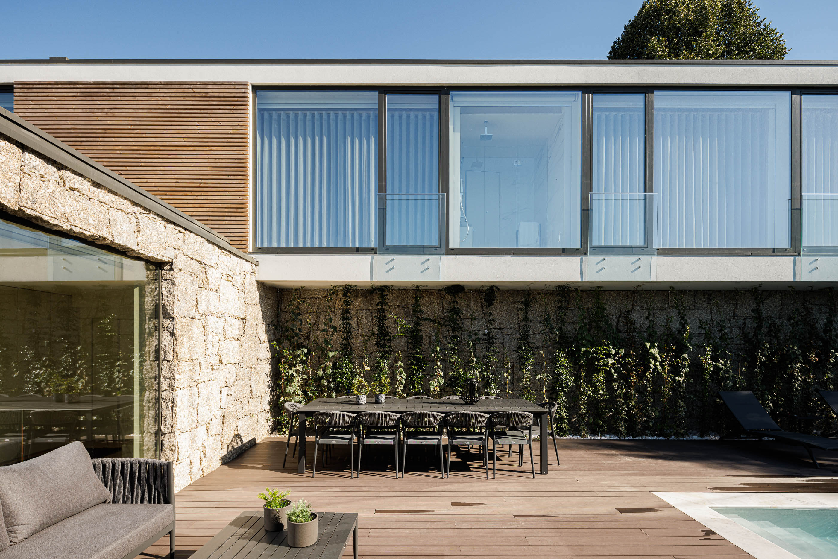 Exterior: Zona de refeições exterior com mesa e cadeiras pretas, piscina e fachada em pedra. — CASA [EN]COSTA arquitetura Hous3 em Guimarães