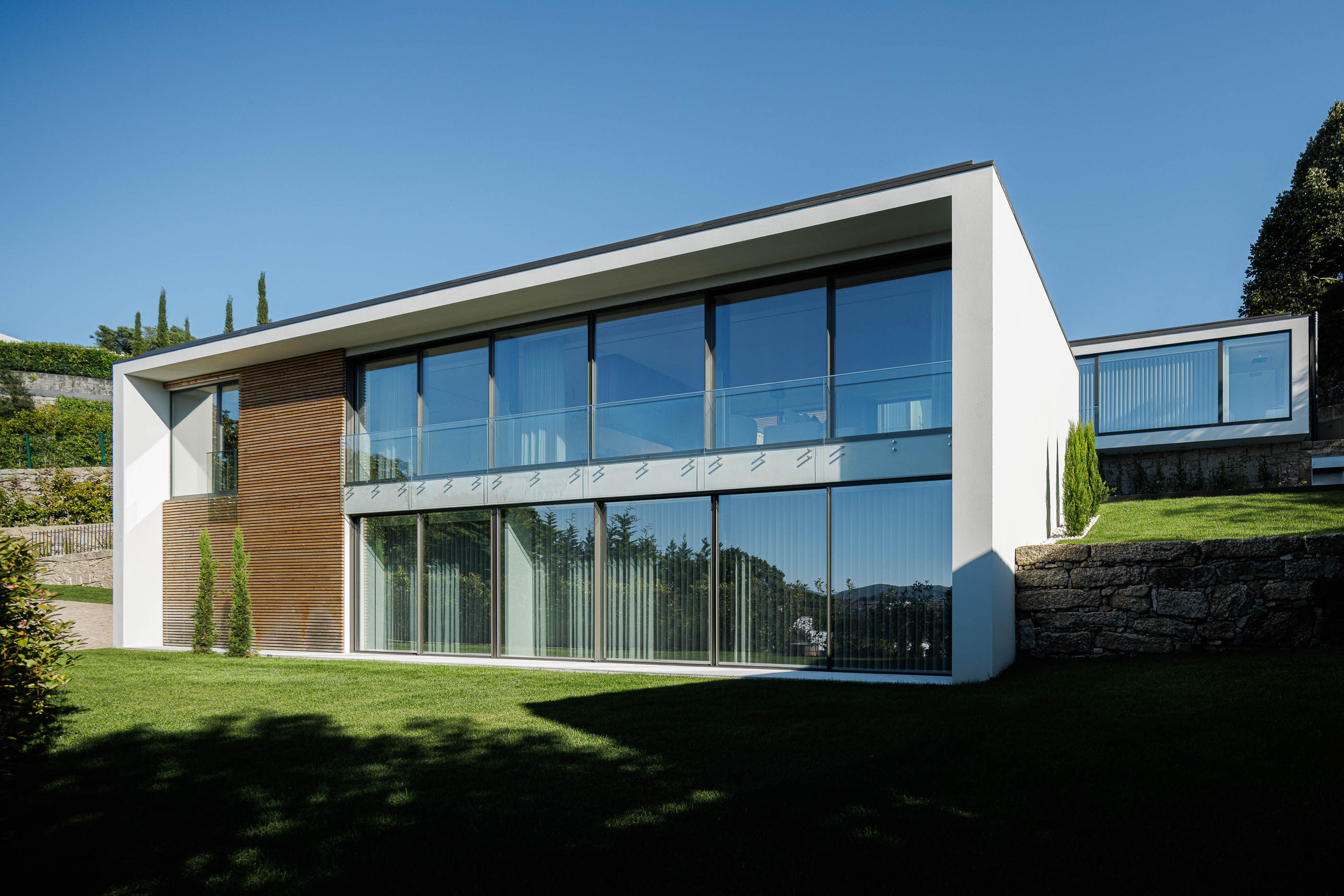Exterior: Vista geral da casa com fachada branca, detalhes em madeira e jardim relvado. — CASA [EN]COSTA arquitetura Hous3 em Guimarães