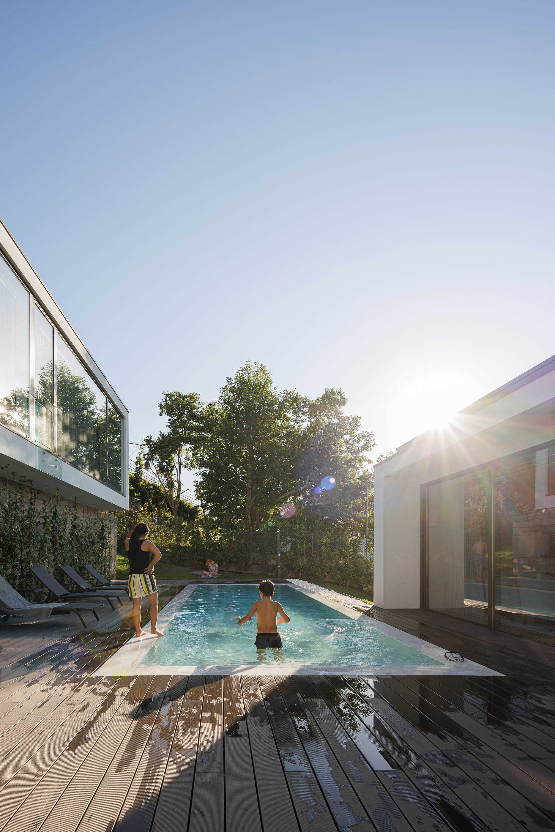 Exterior: Piscina com deck em madeira e vista para a casa, com espreguiçadeiras e vegetação. — CASA [EN]COSTA arquitetura Hous3 em Guimarães