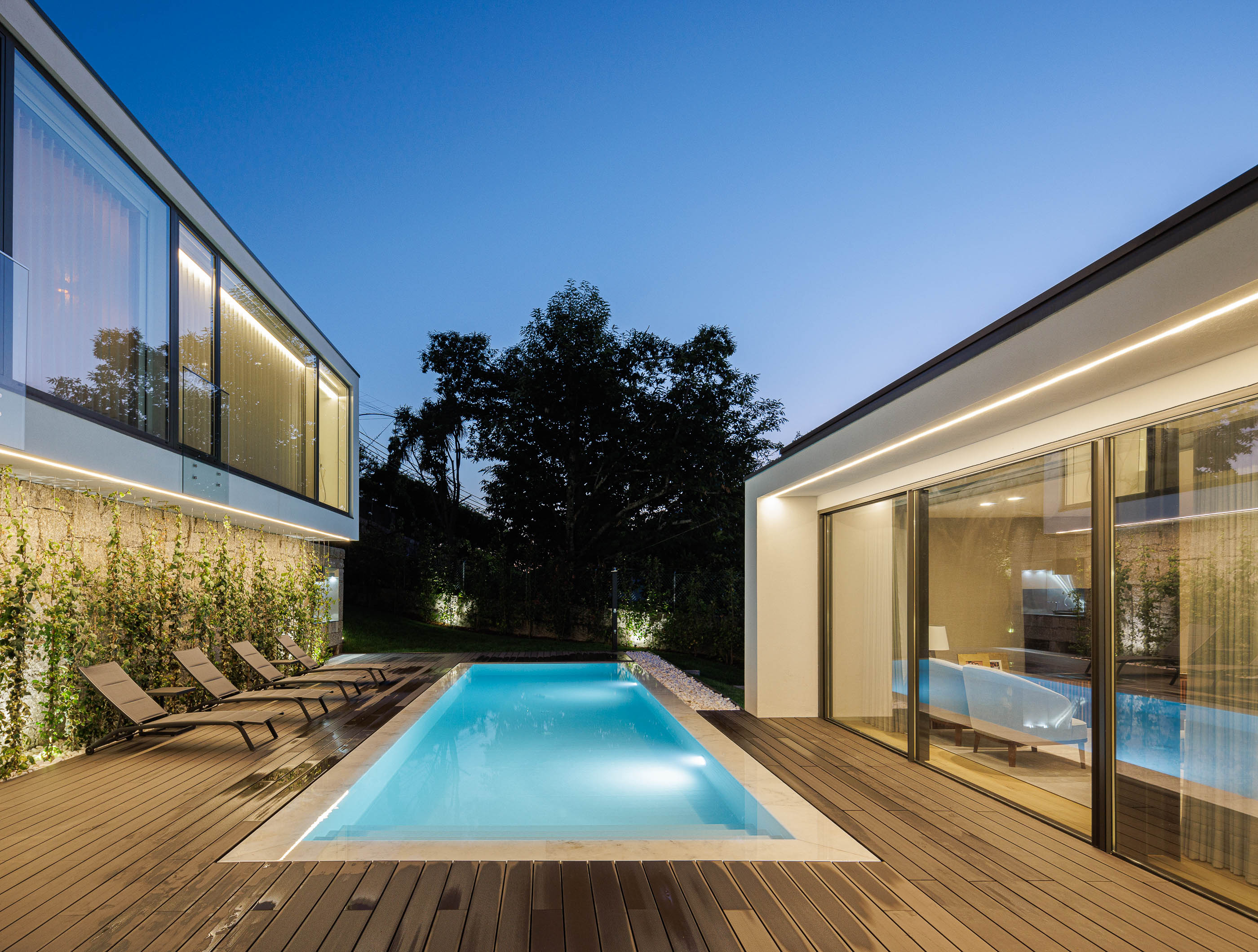Exterior: Piscina iluminada com deck em madeira, espreguiçadeiras e fachada em pedra. — CASA [EN]COSTA arquitetura Hous3 em Guimarães