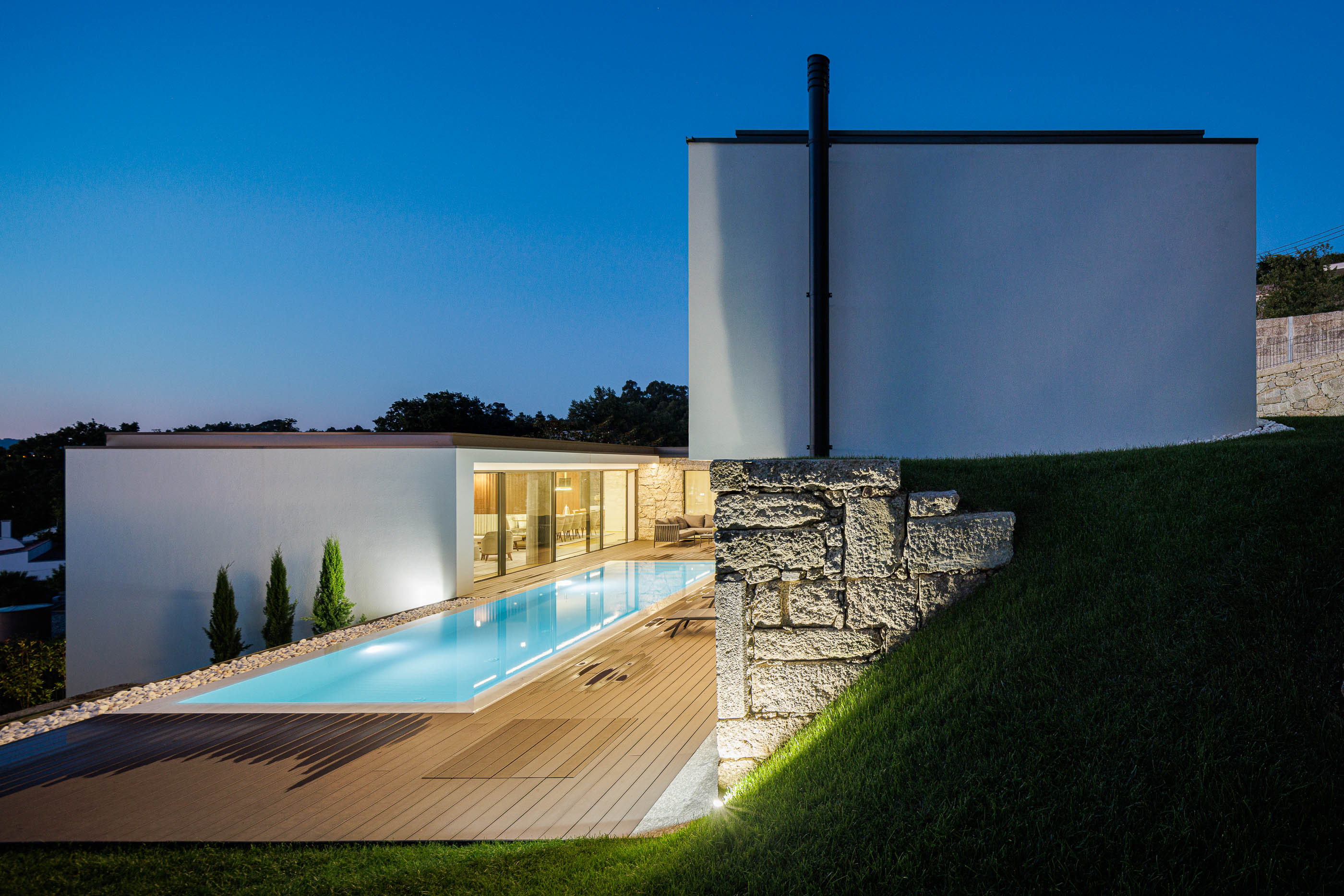 Exterior: Vista noturna da casa com piscina iluminada, deck em madeira e jardim relvado. — CASA [EN]COSTA arquitetura Hous3 em Guimarães