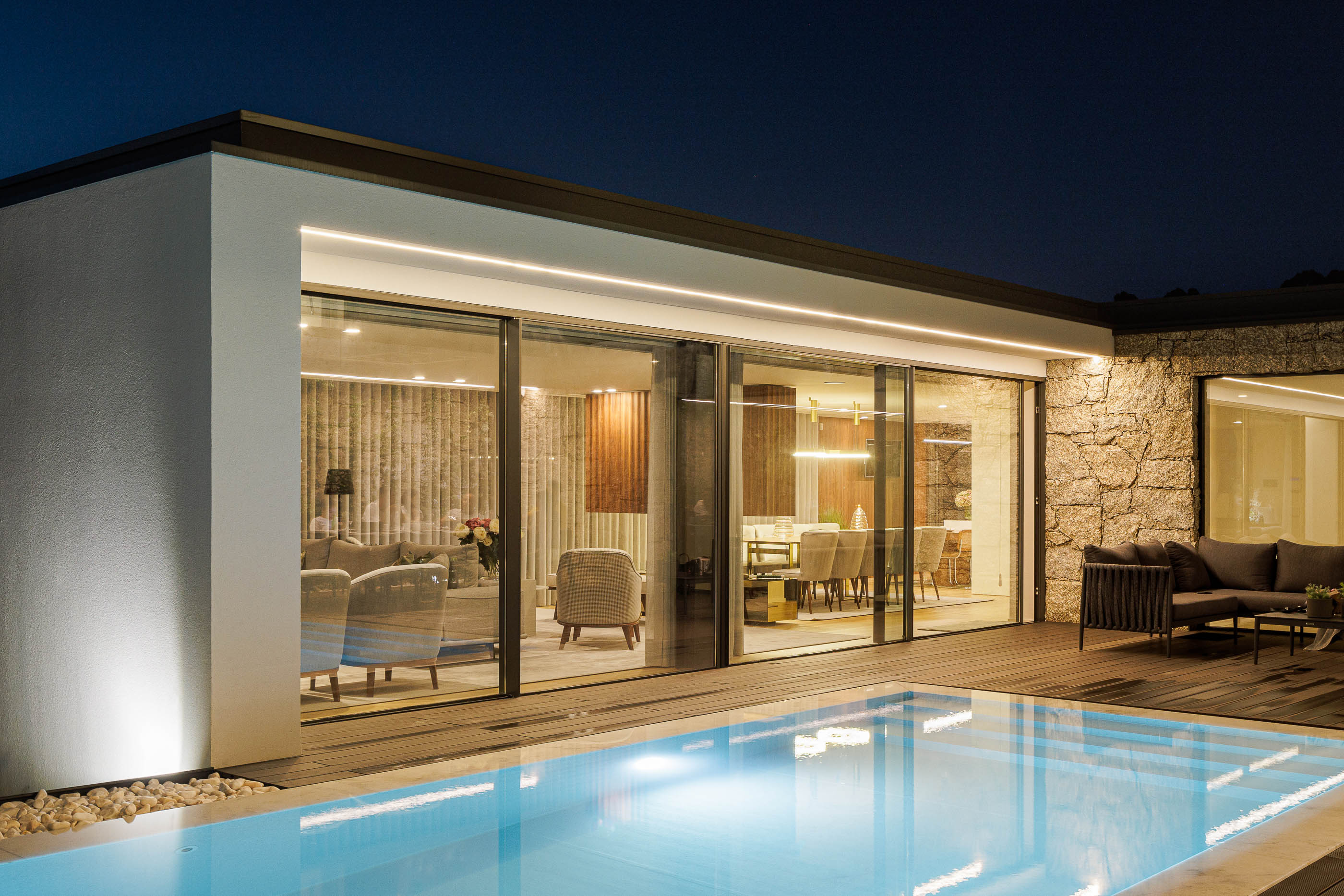 Exterior: Vista noturna da piscina com deck em madeira e janelas iluminadas da sala de estar. — CASA [EN]COSTA arquitetura Hous3 em Guimarães
