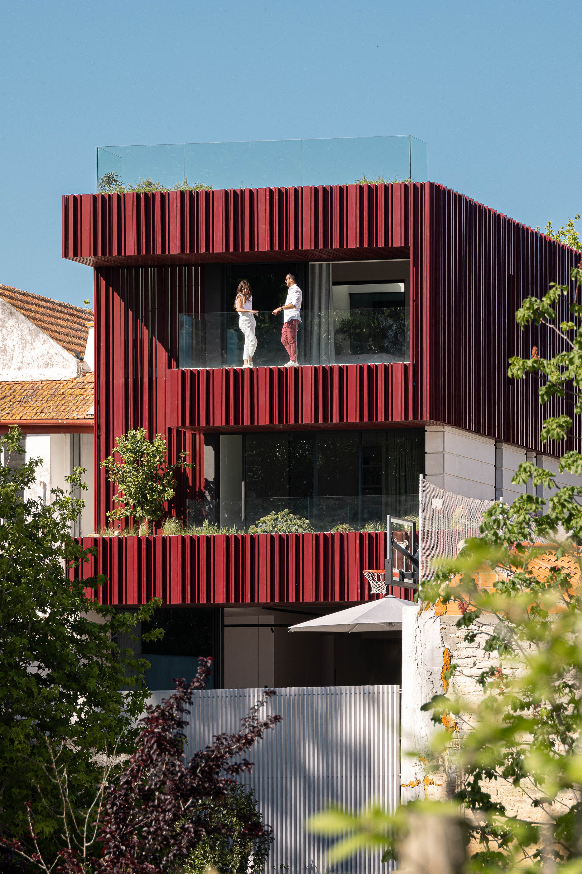 Fachada: Fachada com revestimento vertical vermelho, varandas com vegetação e pessoas. — Casa do Parque arquitetura Paulo Martins Arquitectura e Design em Aveiro