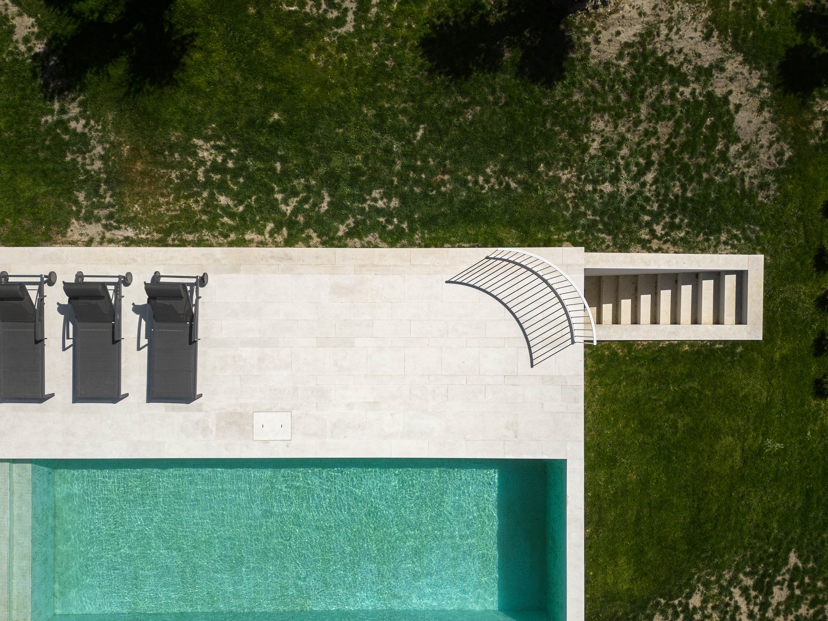 Piscina: Vista superior da piscina retangular com espreguiçadeiras pretas e escadas de acesso, pavimento em pedra clara e relvado. — Casa na Romeira arquitetura dp arquitectos em Santarém