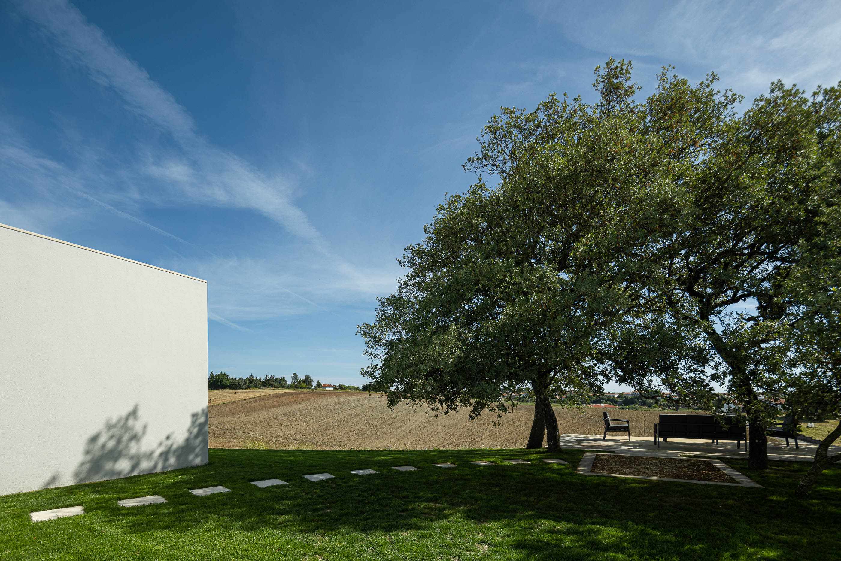 Jardim: Zona de estar exterior com sofás pretos sob a sombra de uma árvore, pavimento em relva com passadeiras de pedra. — Casa na Romeira arquitetura dp arquitectos em Santarém