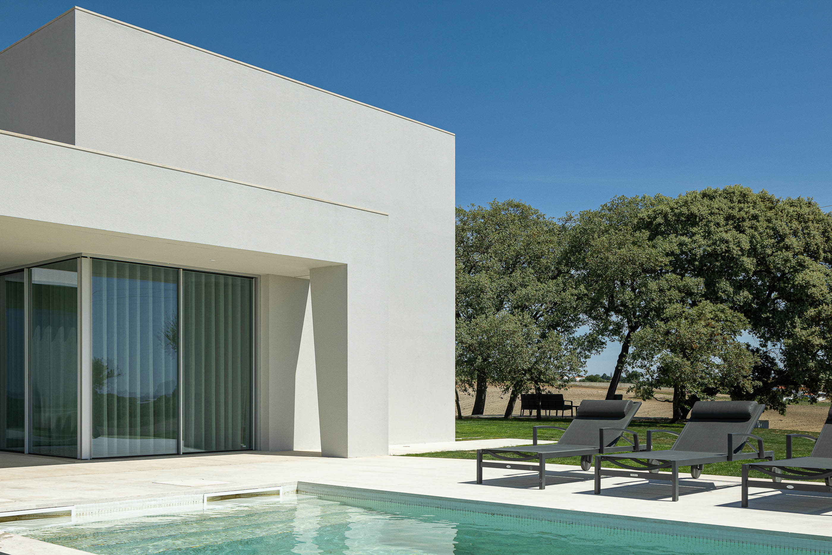 Piscina: Vista da piscina e zona de espreguiçadeiras, com grandes janelas de vidro e alpendre branco. — Casa na Romeira arquitetura dp arquitectos em Santarém