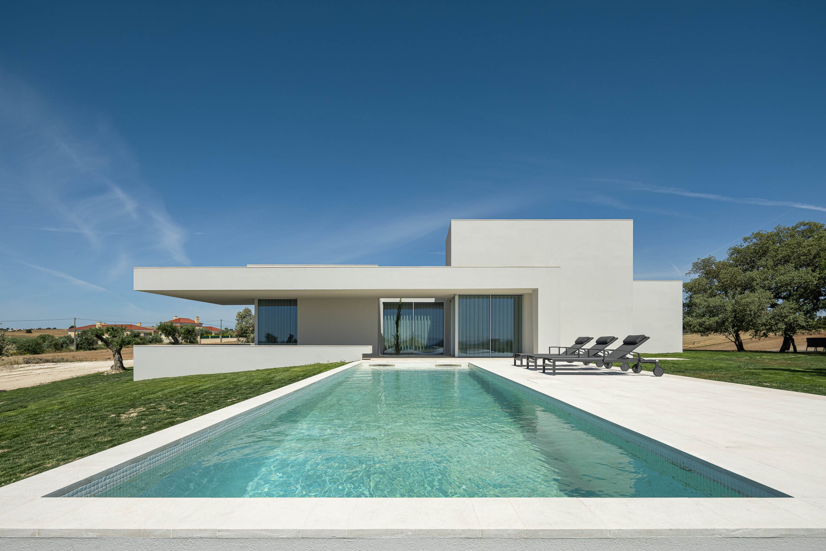 Piscina: Fachada da casa com piscina retangular em primeiro plano, espreguiçadeiras e alpendre. — Casa na Romeira arquitetura dp arquitectos em Santarém