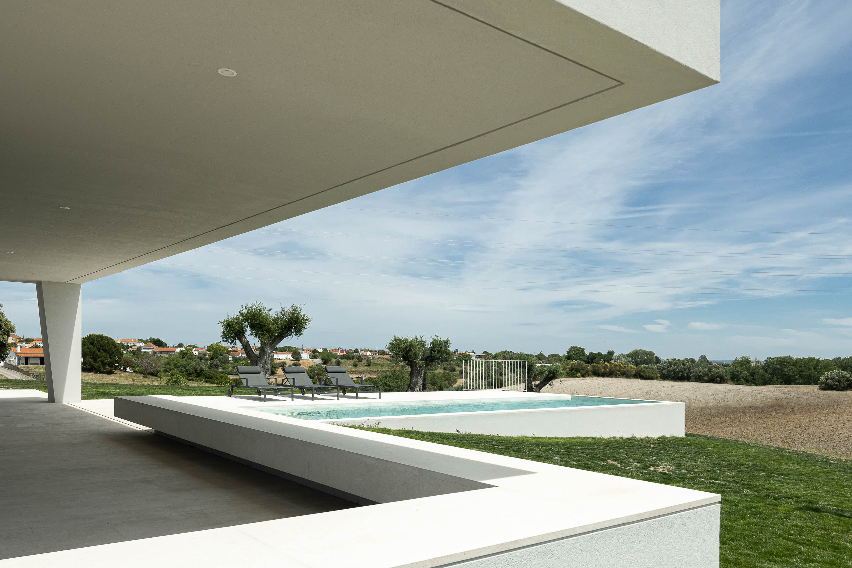 Piscina: Alpendre com vista para a piscina, espreguiçadeiras e paisagem rural. Pavimento em pedra clara. — Casa na Romeira arquitetura dp arquitectos em Santarém