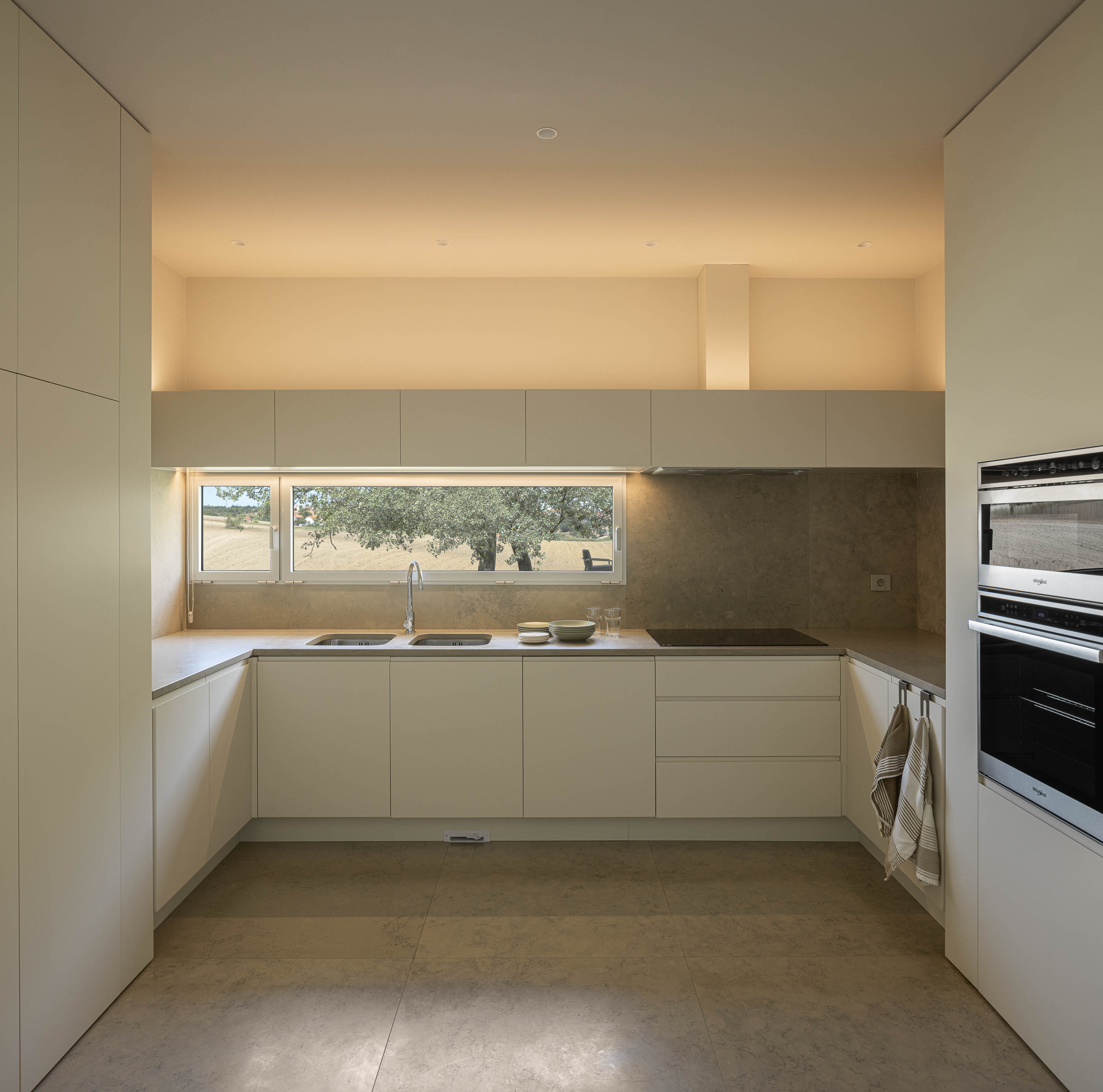 Corredor: Corredor comprido com armários brancos embutidos, candeeiro suspenso de madeira e pavimento cinzento. — Casa na Romeira arquitetura dp arquitectos em Santarém