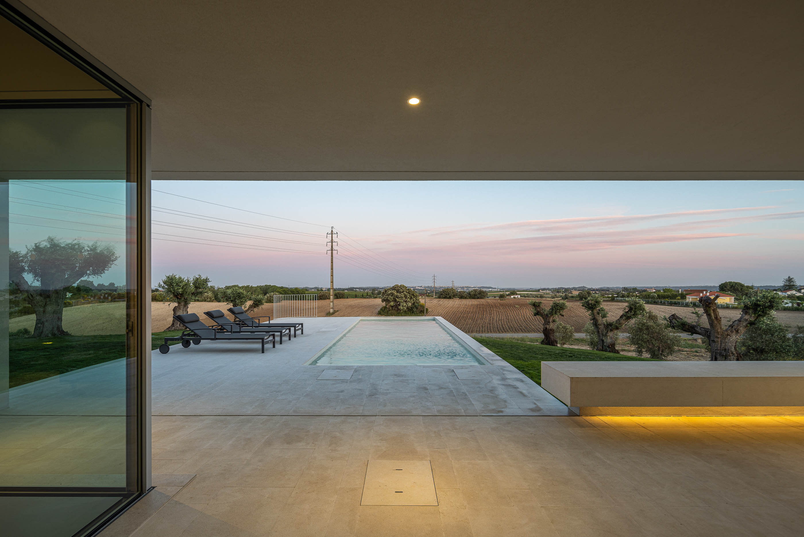 Piscina: Vista da casa branca com piscina, espreguiçadeiras e iluminação exterior ao entardecer. — Casa na Romeira arquitetura dp arquitectos em Santarém