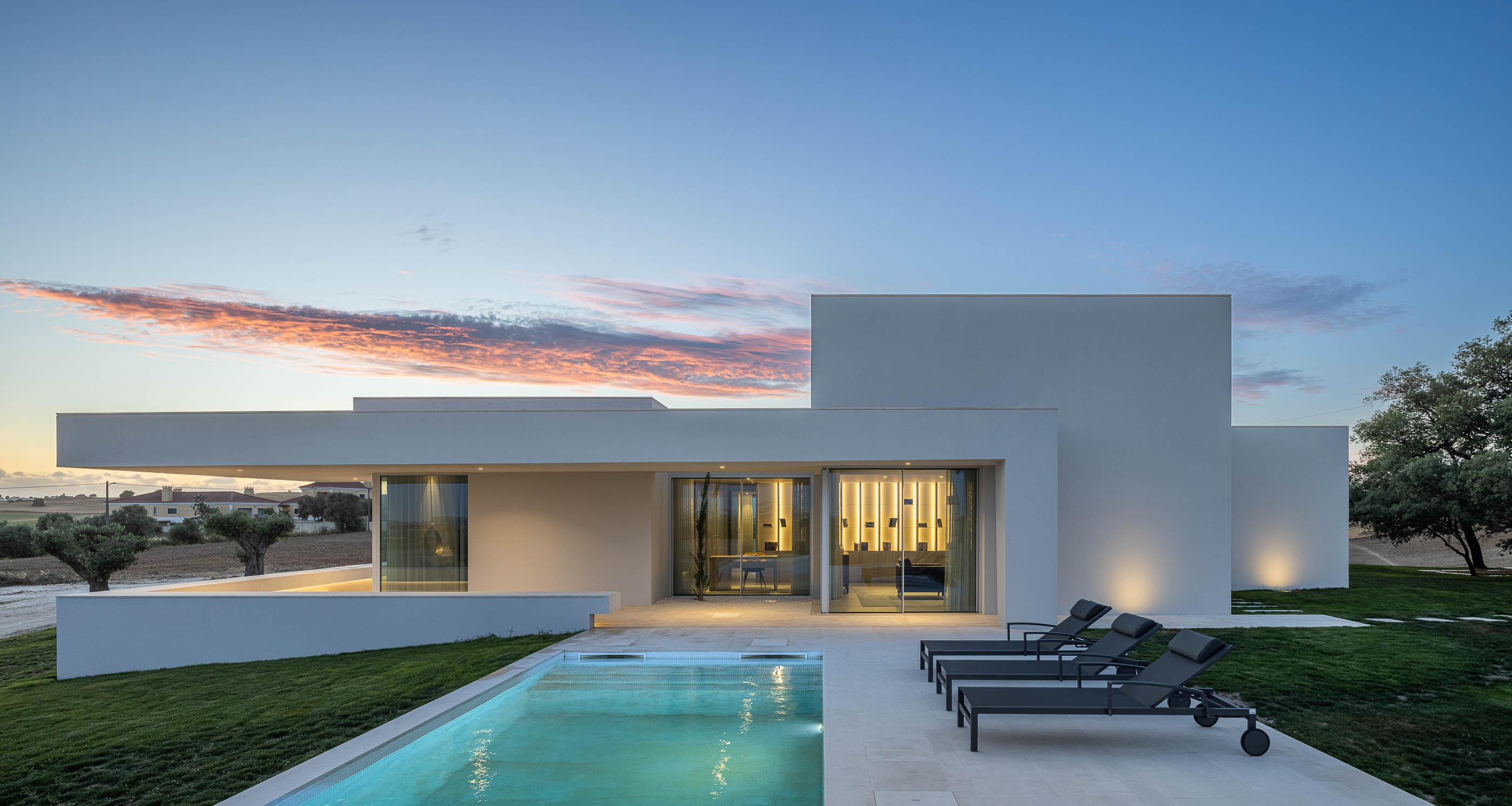 Piscina: Vista da casa branca com piscina, zona de estar exterior e iluminação ao entardecer. — Casa na Romeira arquitetura dp arquitectos em Santarém