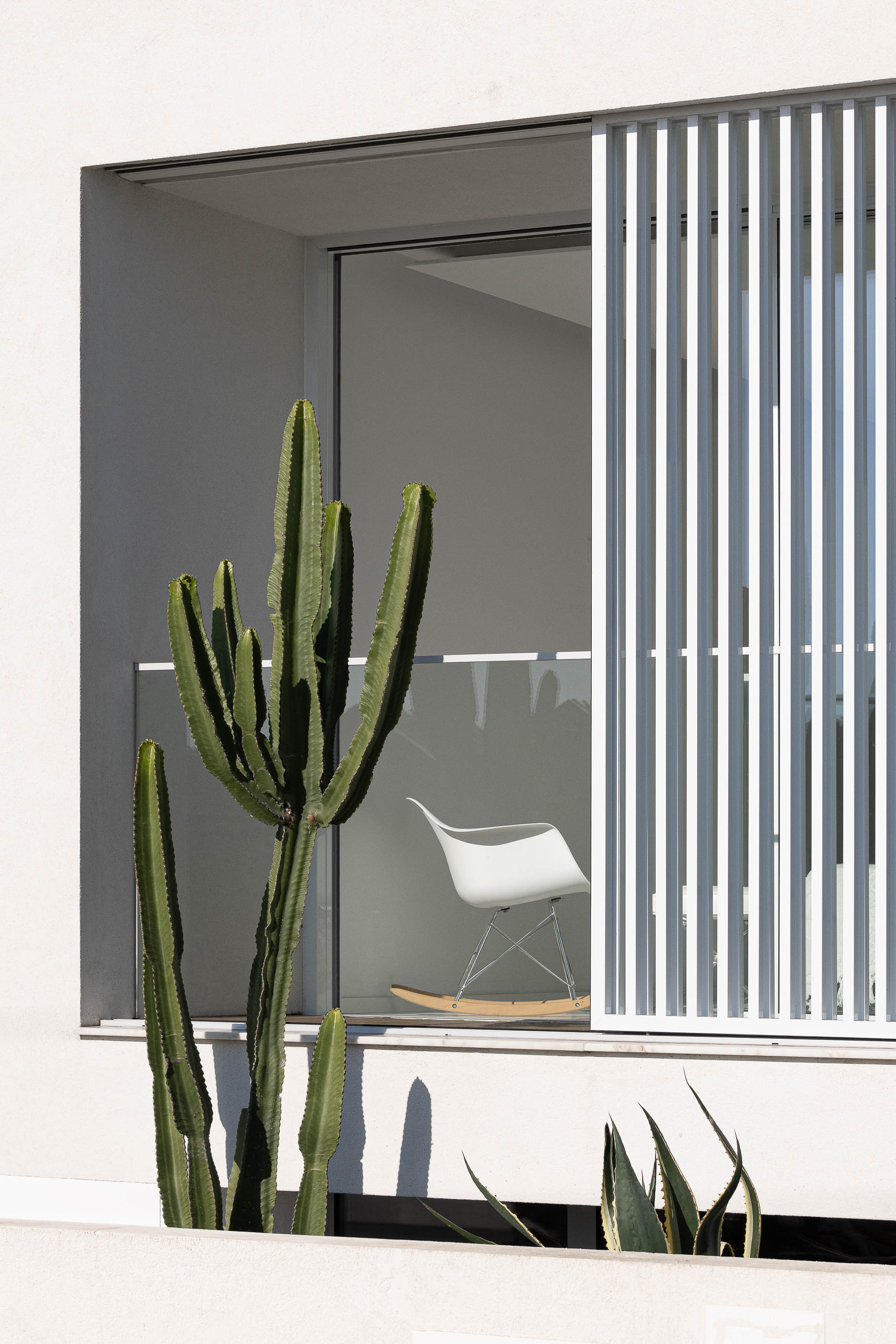 Varanda: Cadeira de baloiço branca em varanda com pavimento de madeira, protegida por brises verticais brancos e cactos. — Casa Cor de Sal arquitetura Rómulo Neto Arquitectos em Ílhavo