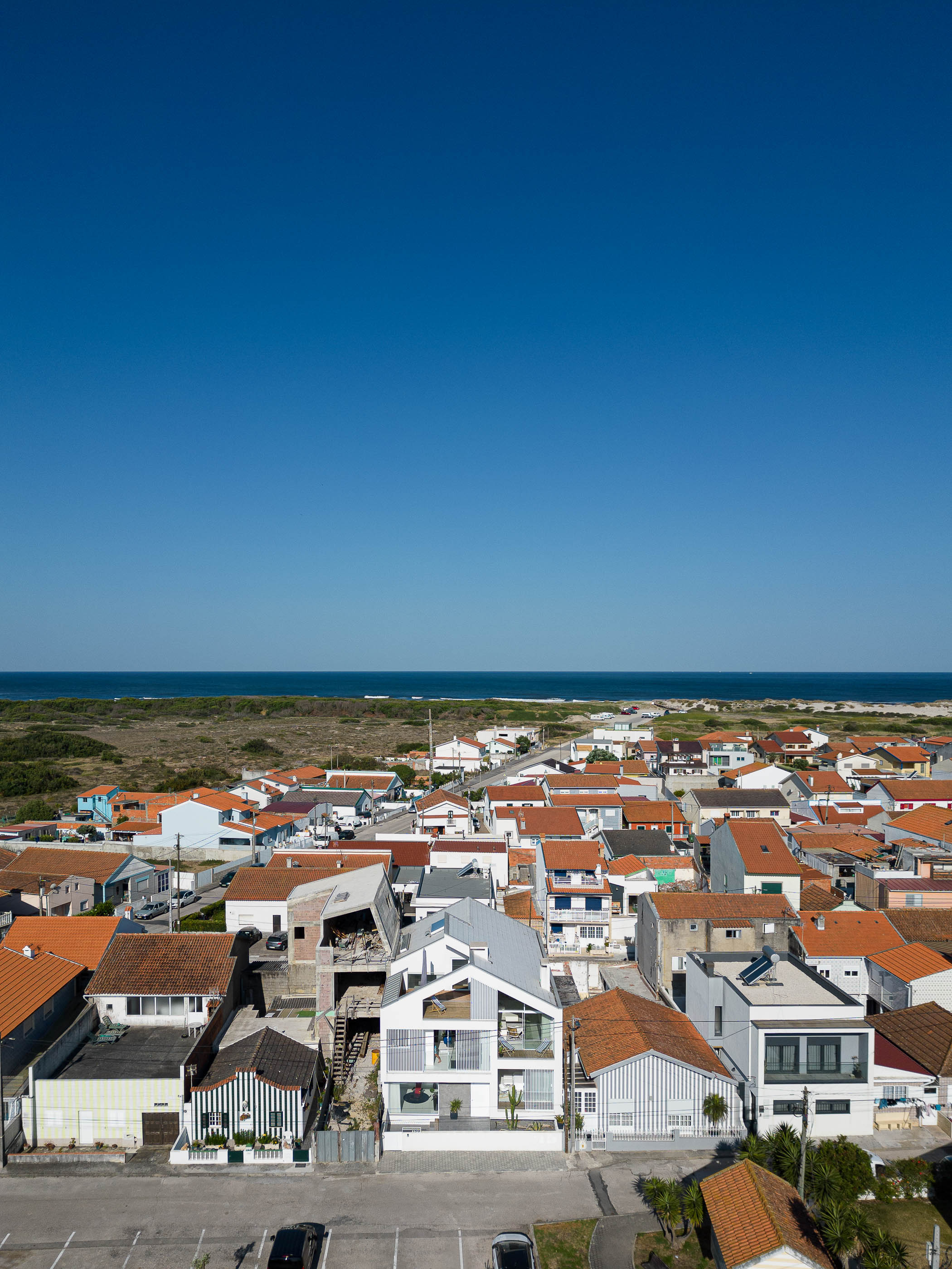 Exterior: Vista aérea da casa integrada na malha urbana, com telhados de cor laranja e o oceano ao fundo. — Casa Cor de Sal arquitetura Rómulo Neto Arquitectos em Ílhavo
