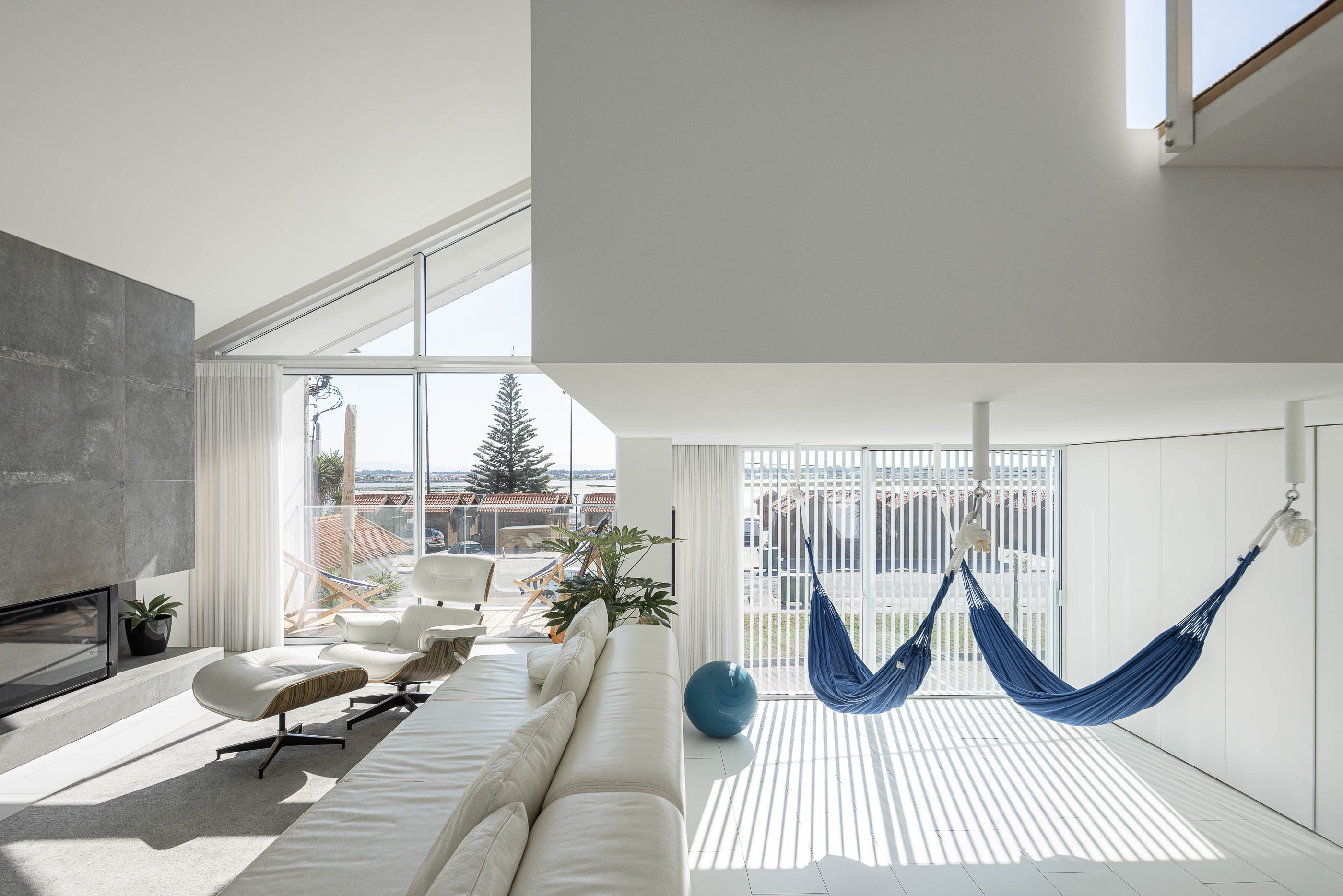 Sala de estar: Espaço amplo com sofá branco, lareira revestida a pedra cinzenta e rede azul suspensa. — Casa Cor de Sal arquitetura Rómulo Neto Arquitectos em Ílhavo