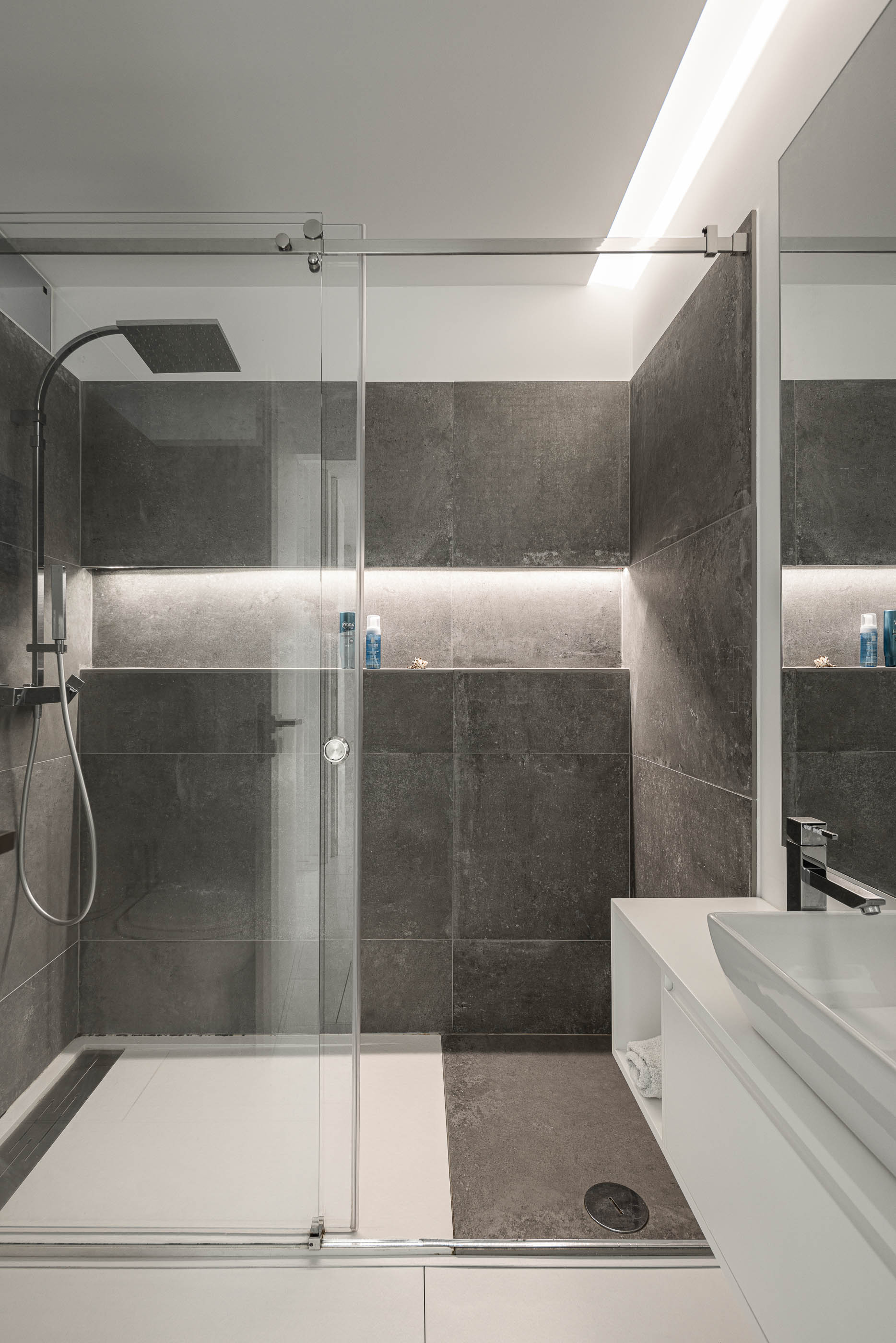 Casa de banho: Cabine de duche com revestimento cinzento, lavatório branco e iluminação embutida. — Casa Cor de Sal arquitetura Rómulo Neto Arquitectos em Ílhavo