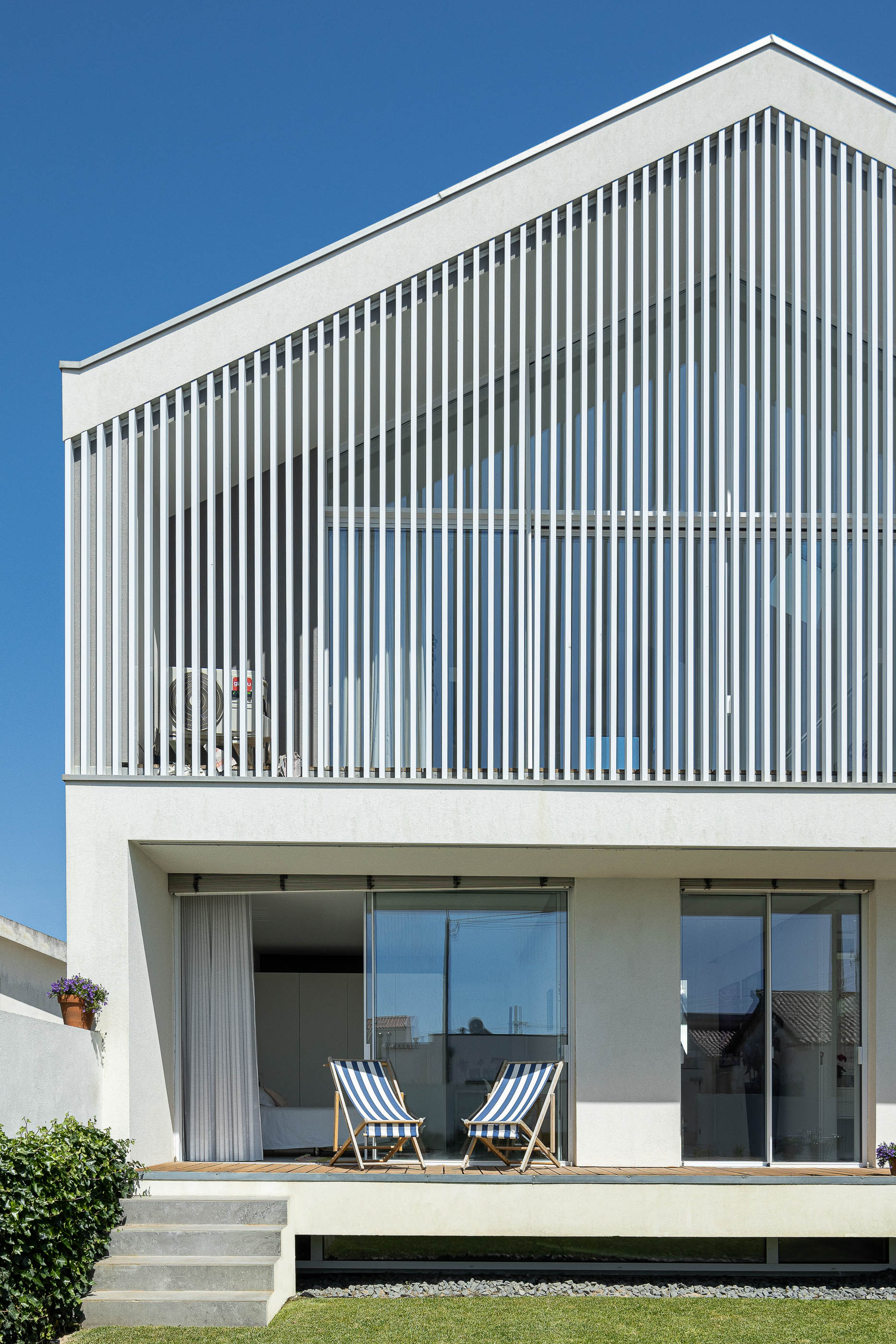 Exterior: Fachada da casa com varanda protegida por brises verticais, cadeiras de praia e jardim relvado. — Casa Cor de Sal arquitetura Rómulo Neto Arquitectos em Ílhavo