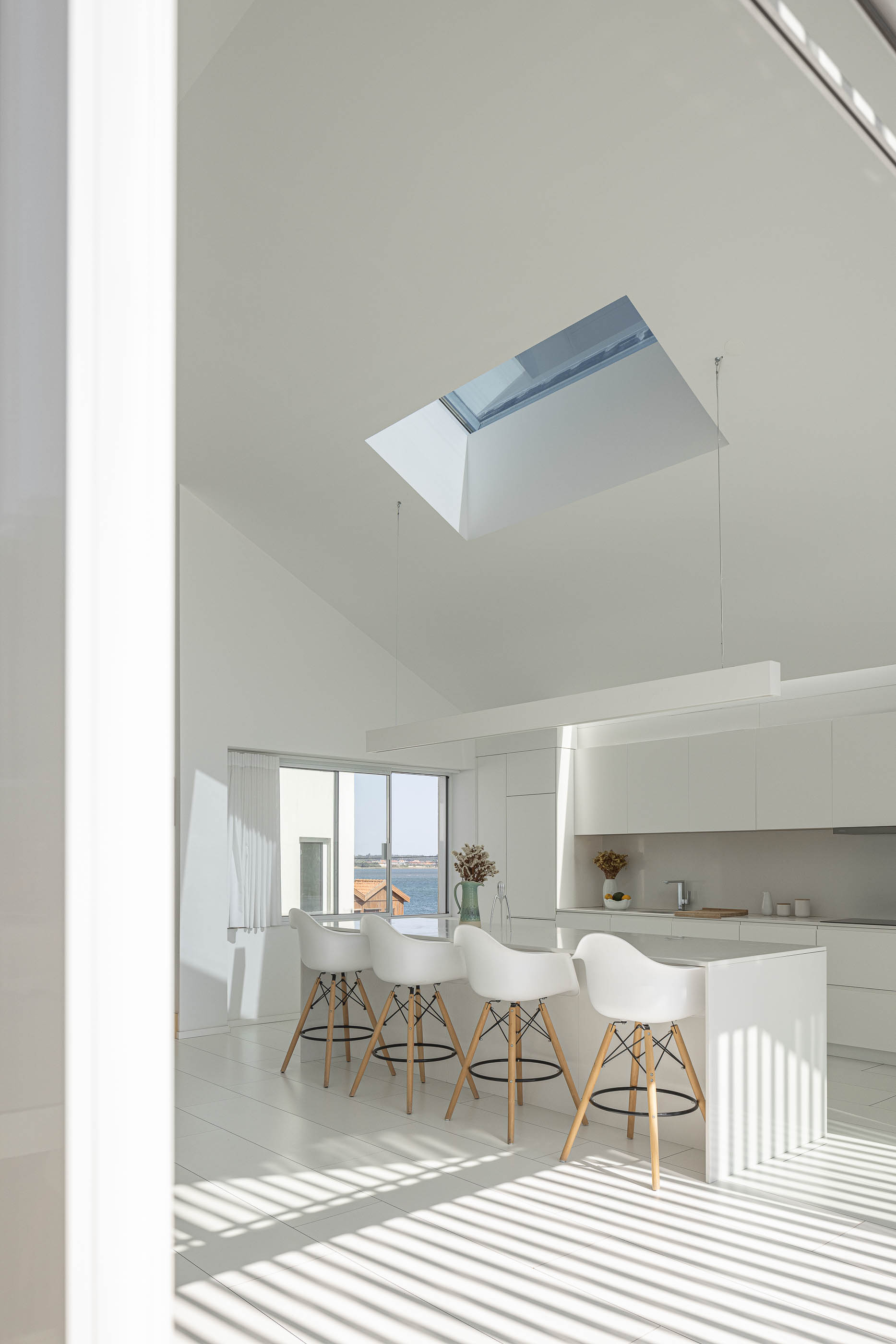 Cozinha: Ilha central com bancos brancos, armários brancos e claraboia no teto inclinado. — Casa Cor de Sal arquitetura Rómulo Neto Arquitectos em Ílhavo