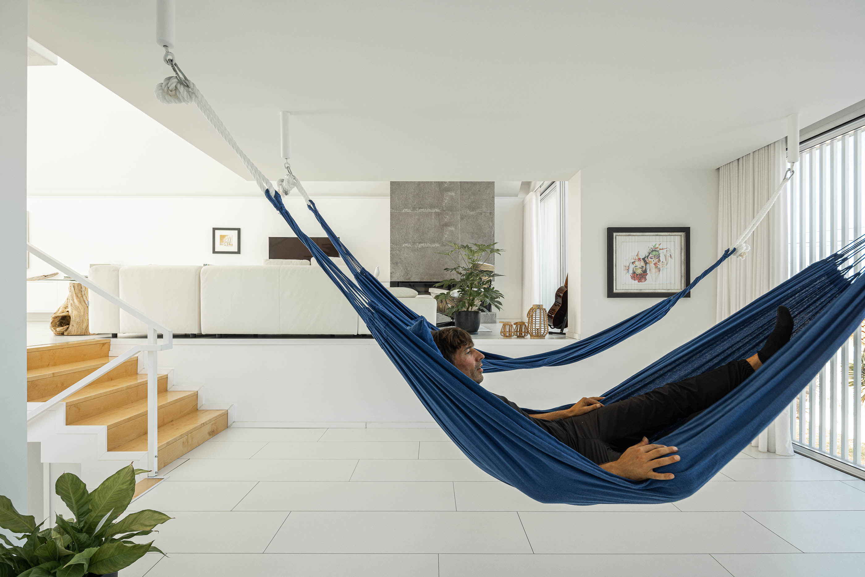 Sala de estar: Homem deitado na rede azul suspensa, com vista para a sala de estar e escadaria. — Casa Cor de Sal arquitetura Rómulo Neto Arquitectos em Ílhavo