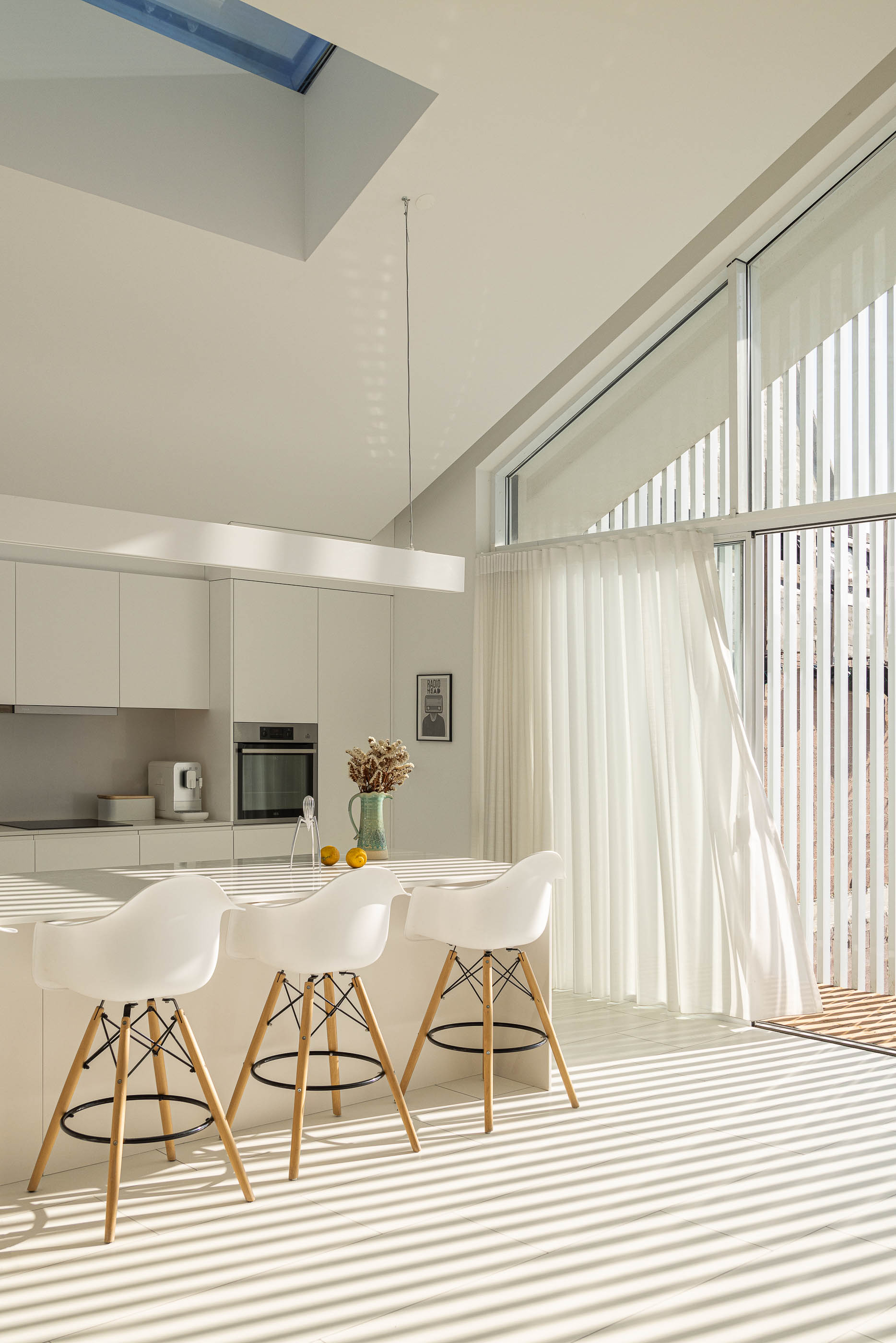 Cozinha: Ilha central com bancos brancos, armários brancos e vista para o exterior através da janela. — Casa Cor de Sal arquitetura Rómulo Neto Arquitectos em Ílhavo