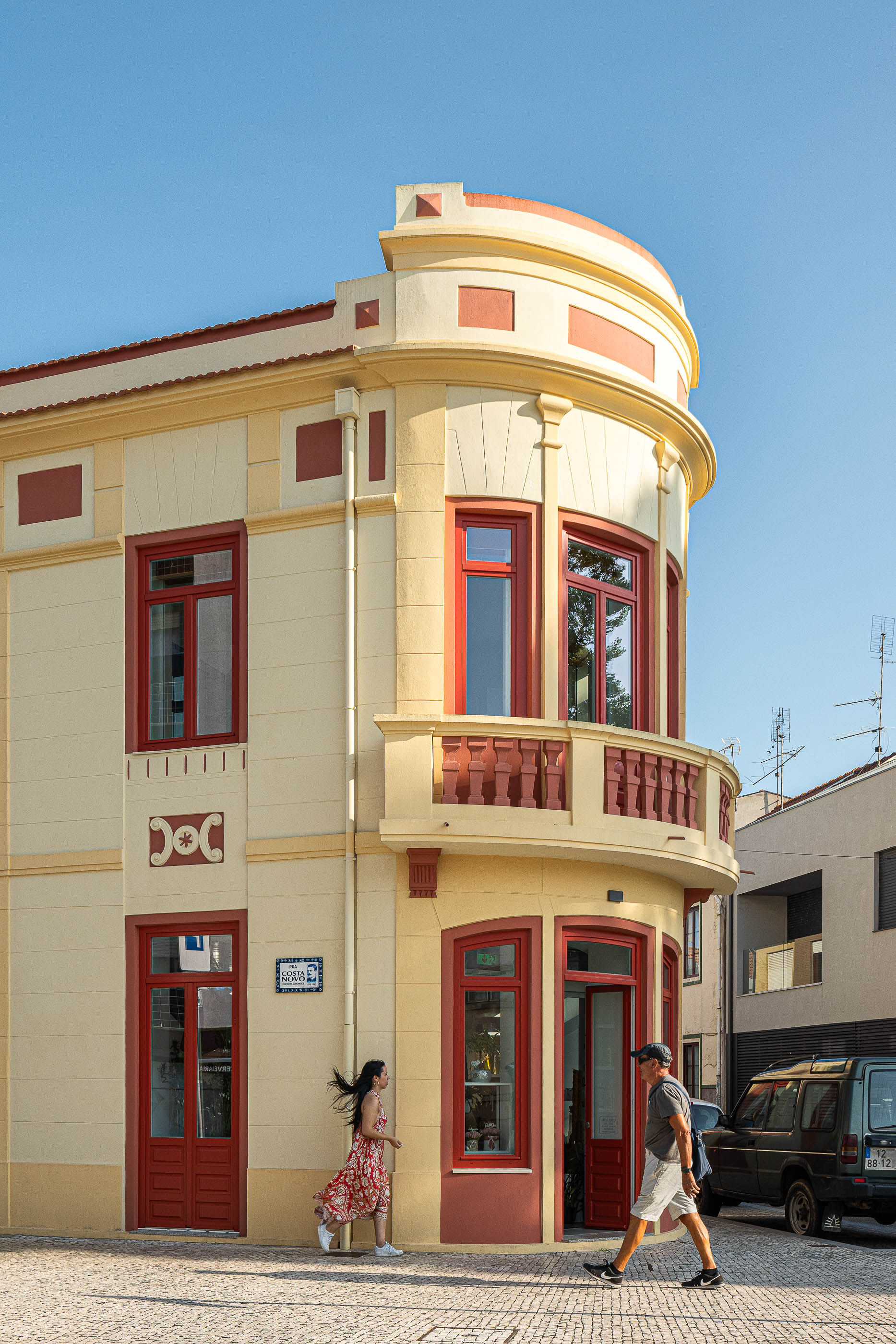 Fachada: Edifício de esquina com detalhes clássicos, em tons creme e vermelho, com varanda curva e pavimento em calçada. — A'mar arquitetura REM'A Arquitectos em Póvoa de Varzim