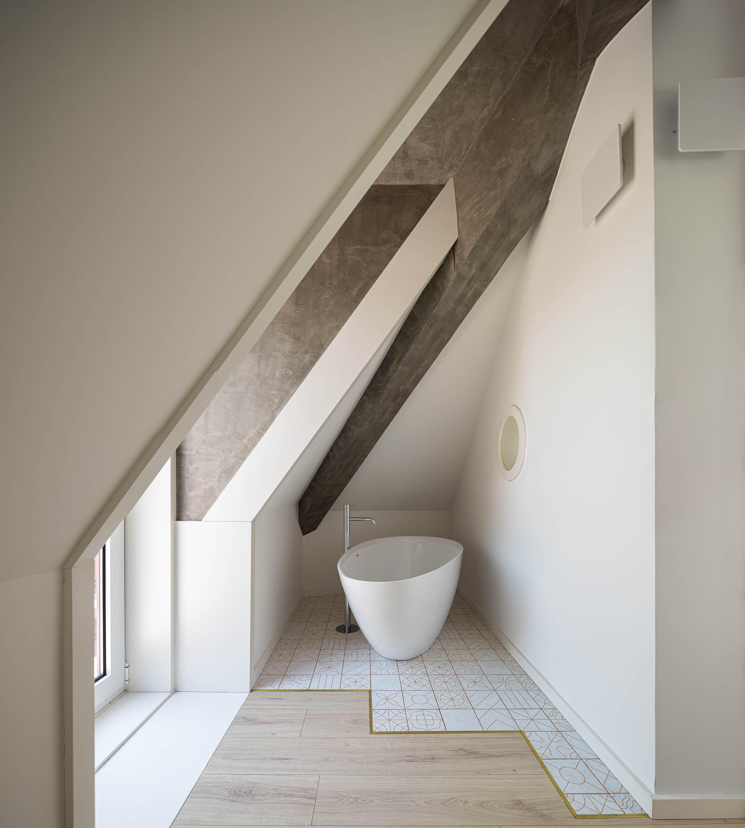 Casa de banho: Banheira branca sob teto inclinado, com pavimento cerâmico decorado e janela lateral. — A'mar arquitetura REM'A Arquitectos em Póvoa de Varzim