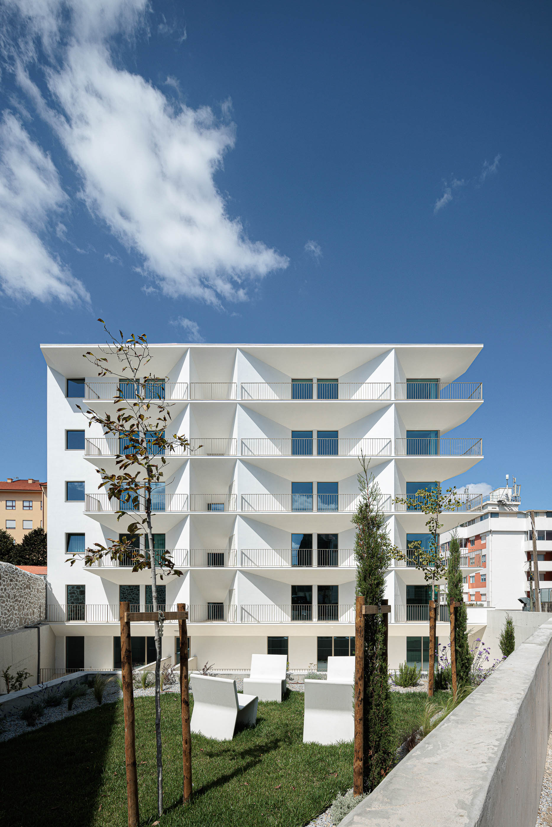 Fachada: Edifício branco com varandas triangulares, guardas metálicas e jardim relvado com árvores jovens. — Varandas de Salgueiros arquitetura Floret Arquitectura em Porto