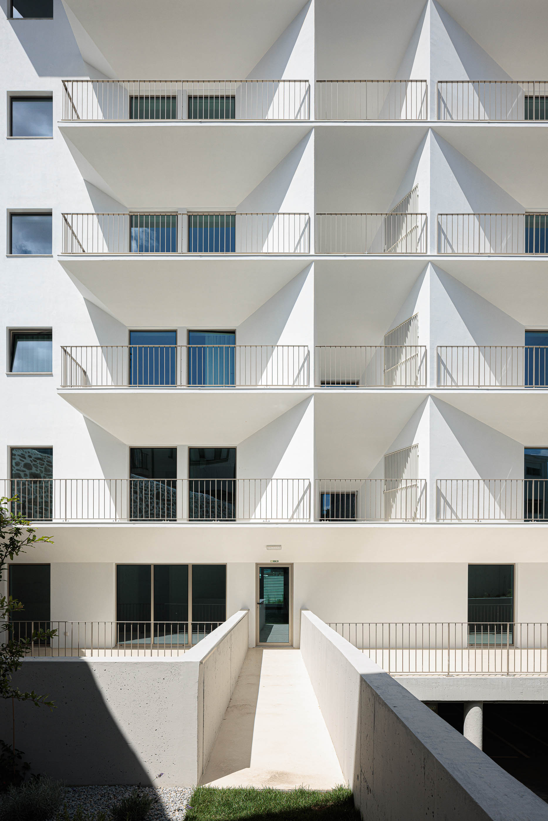 Fachada: Edifício branco com varandas triangulares, acesso pedonal elevado e vegetação. — Varandas de Salgueiros arquitetura Floret Arquitectura em Porto