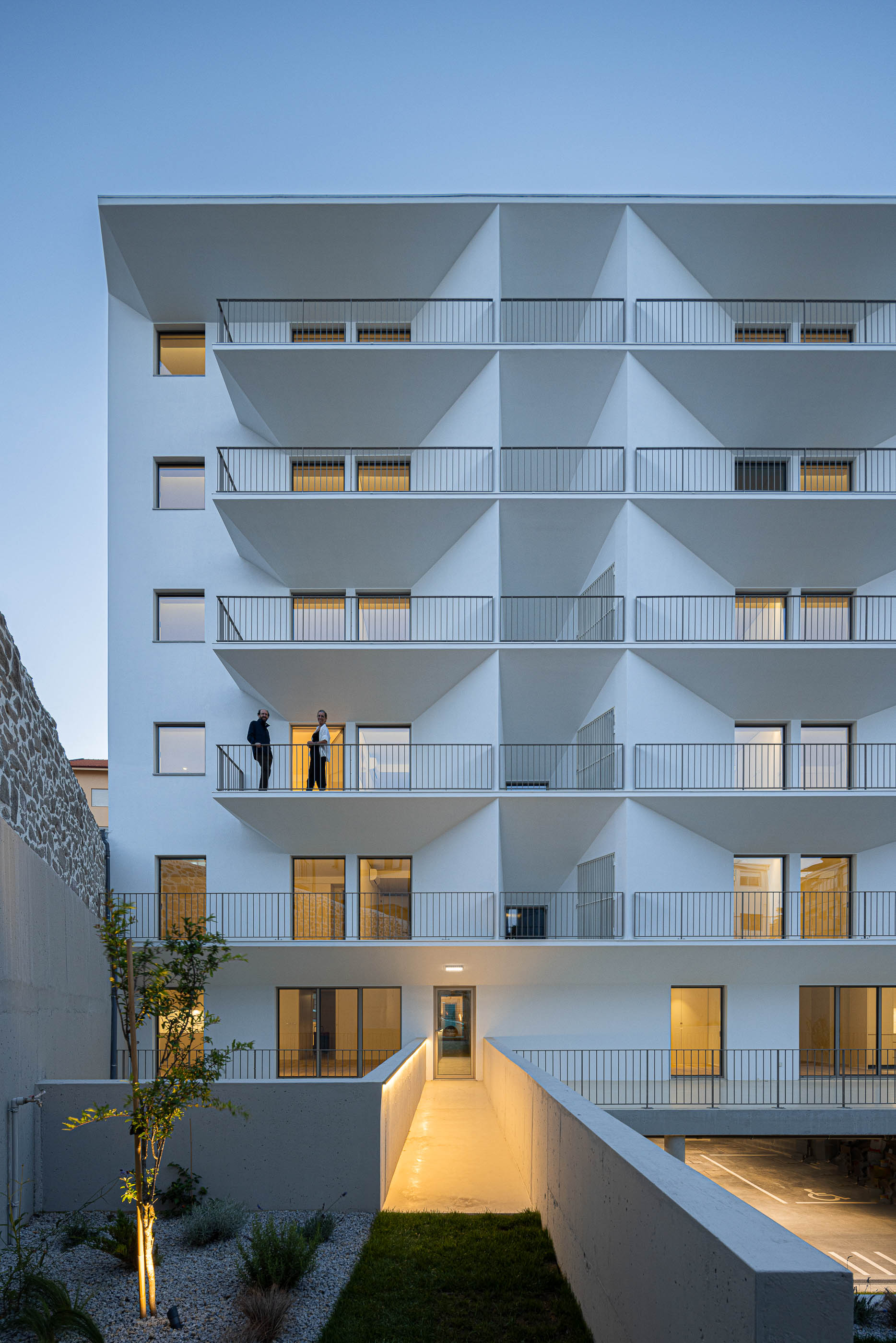 Exterior: Edifício branco com varandas triangulares e acesso pedonal elevado iluminado. — Varandas de Salgueiros arquitetura Floret Arquitectura em Porto