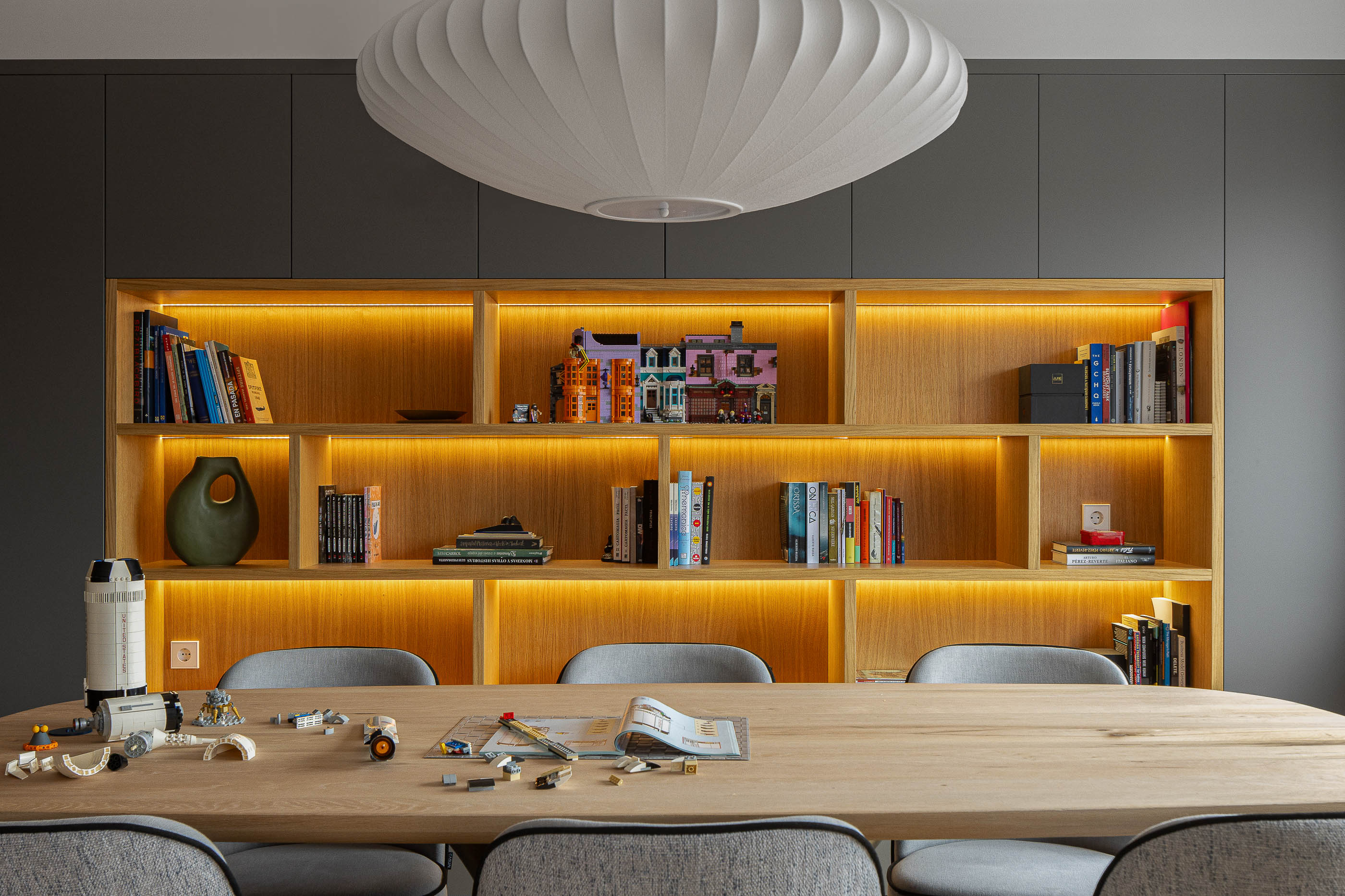 Sala de jantar com mesa de madeira clara, cadeiras cinzentas e estante iluminada com objetos decorativos. — Reforma apartamento CP em Avilés arquitetura David Olmos Arquitectos em Avilés