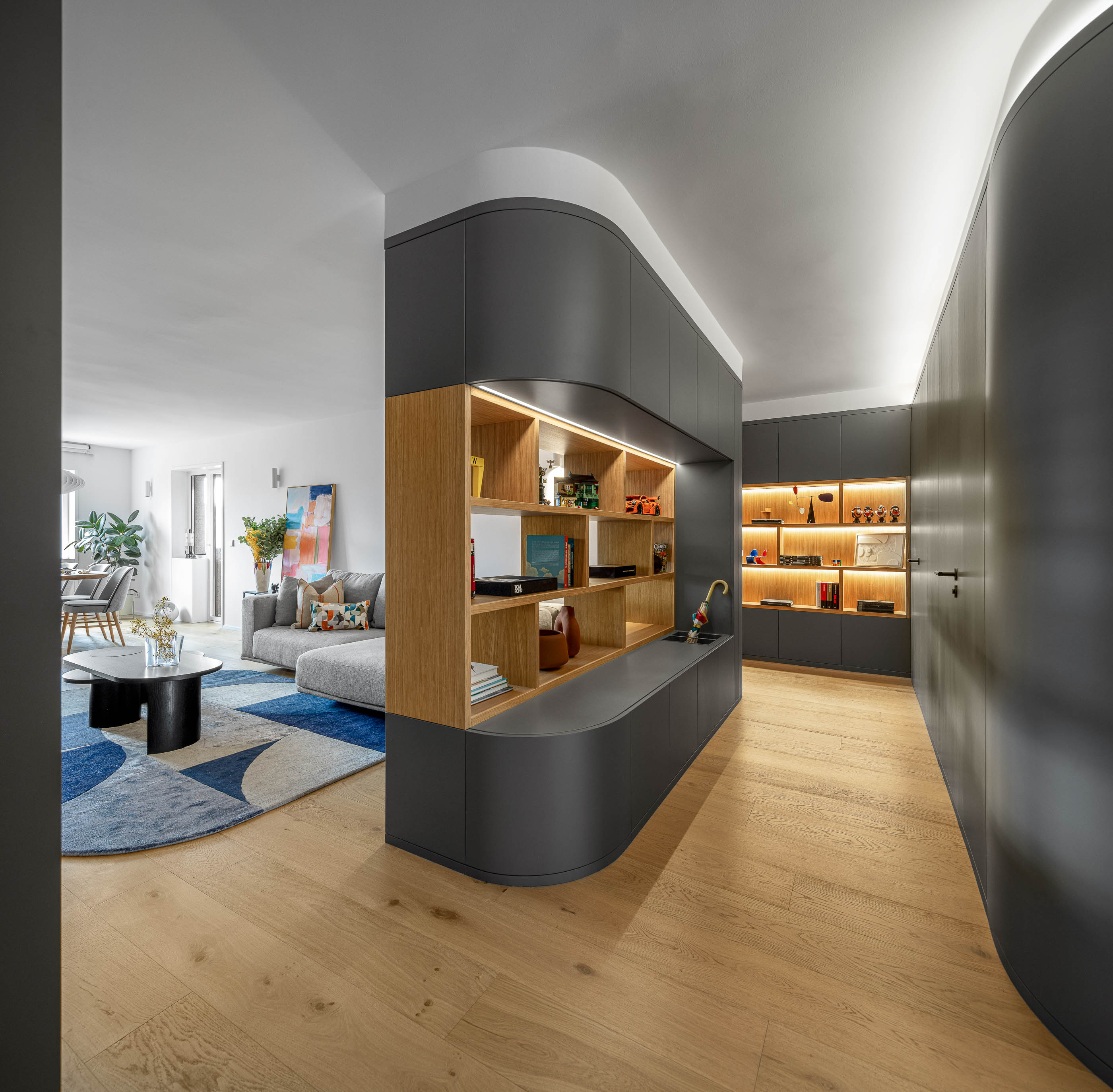 Corredor e sala de estar com móvel cinzento curvo, estantes iluminadas e pavimento em madeira clara. — Reforma apartamento CP em Avilés arquitetura David Olmos Arquitectos em Avilés