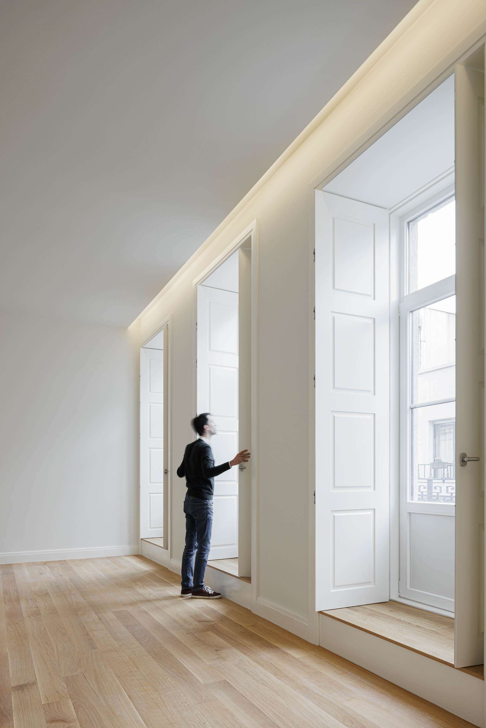 Sala de estar: Homem a abrir uma das portadas brancas, pavimento em madeira clara. — Condomínio das Artes arquitetura MiMool Arquitectura & Interiores em Porto