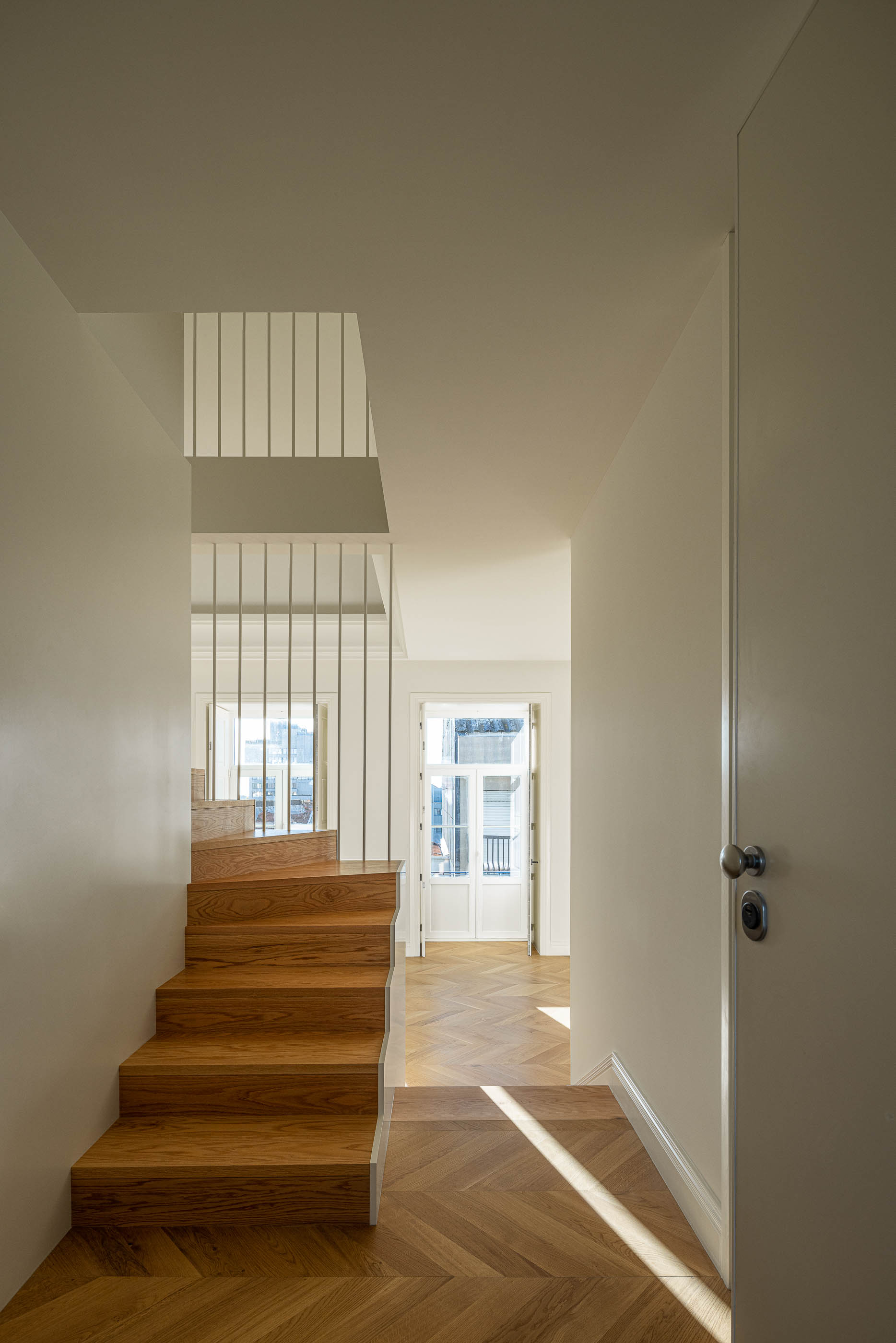 Corredor: Escadas em madeira clara, pavimento em madeira espinha de peixe e porta branca. — Condomínio das Artes arquitetura MiMool Arquitectura & Interiores em Porto