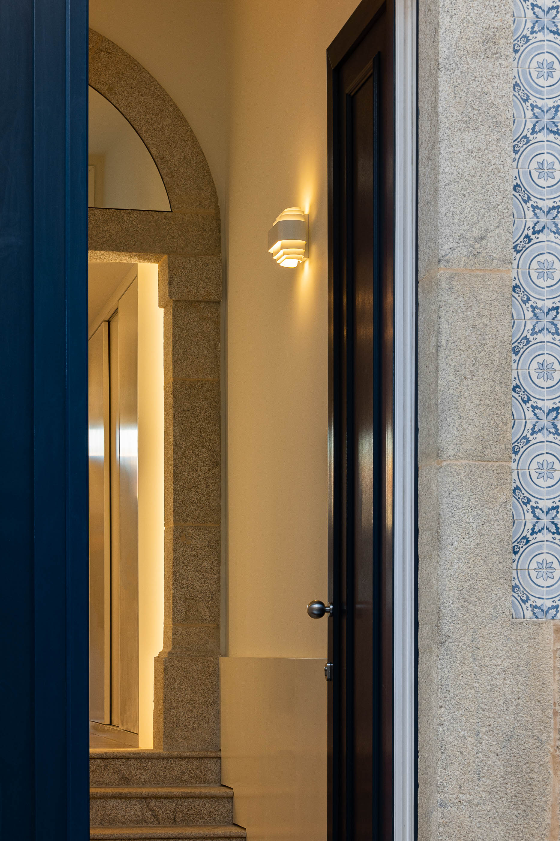 Hall de entrada: Detalhe do candeeiro de parede, azulejos azuis e porta em madeira escura. — Condomínio das Artes arquitetura MiMool Arquitectura & Interiores em Porto