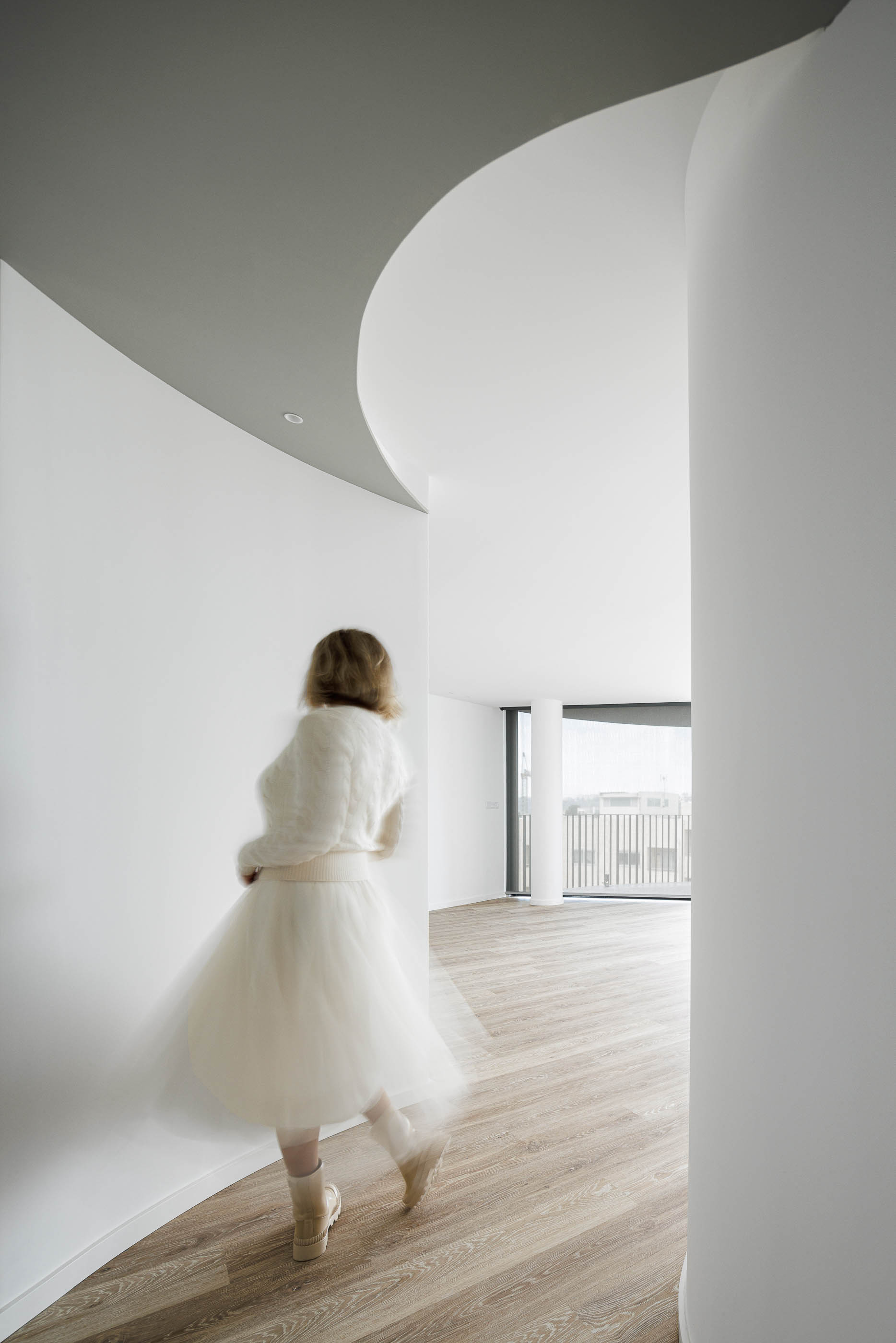 Hall de entrada: Vista do corredor com pavimento em madeira, paredes brancas curvas e teto cinzento com iluminação. — Edifício Sense 100 arquitetura Sandra Micaela Casinha Atelier em Rio Tinto