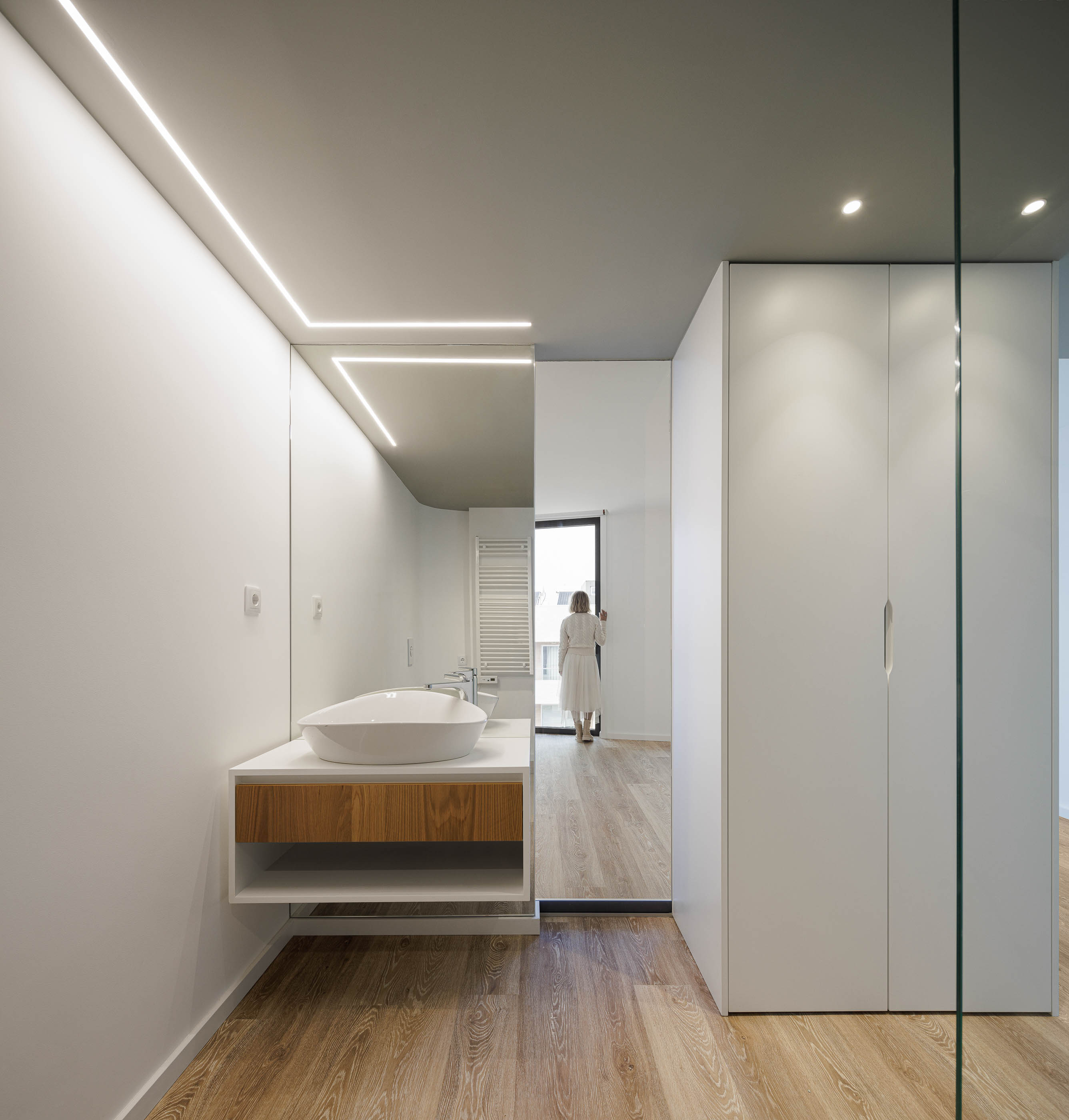 Casa de banho: Lavatório moderno com espelho iluminado, armário em madeira, pavimento em madeira e cabine de duche em vidro. — Edifício Sense 100 arquitetura Sandra Micaela Casinha Atelier em Rio Tinto