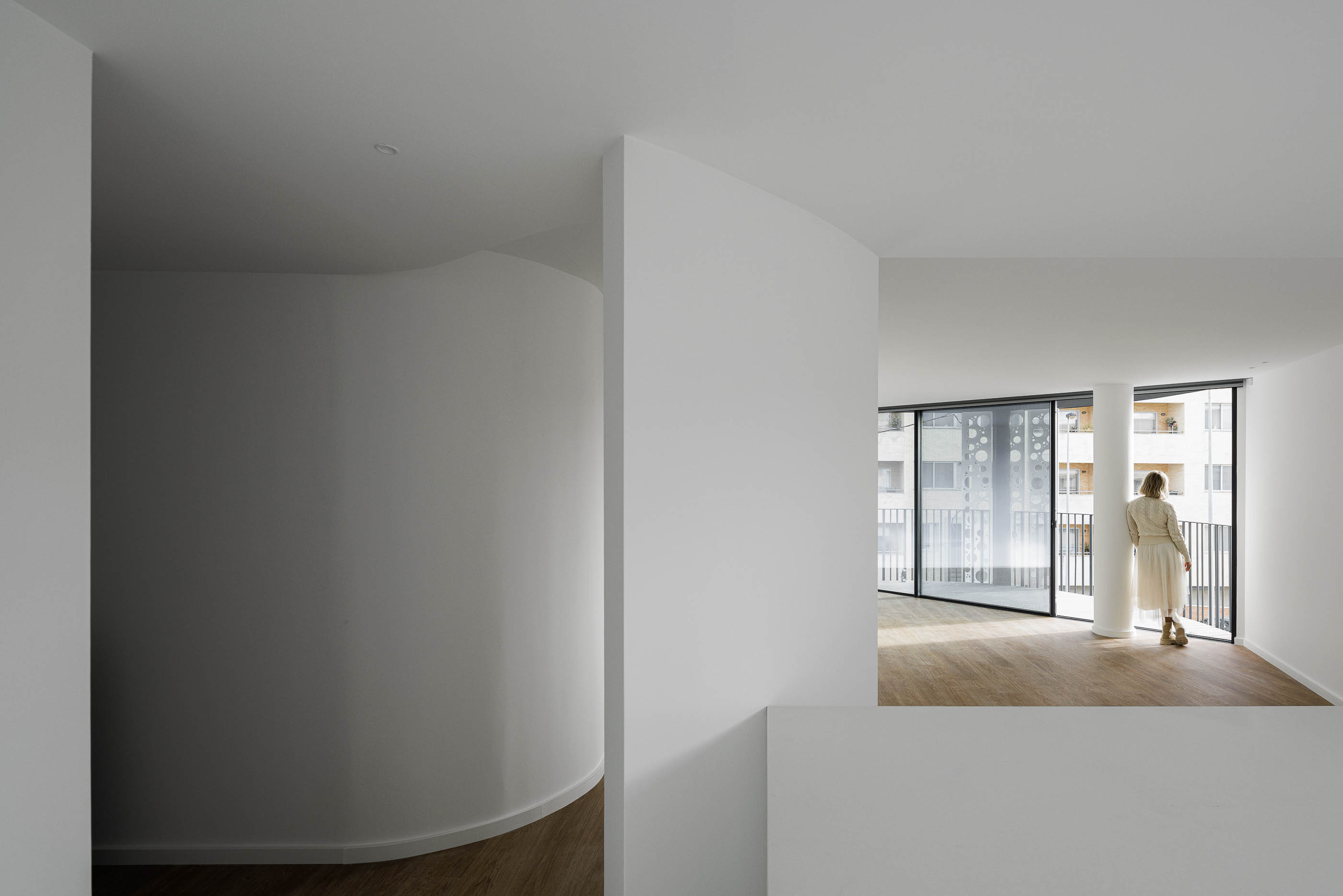 Hall de entrada: Vista do corredor com paredes curvas brancas, pavimento em madeira e acesso à varanda. — Edifício Sense 100 arquitetura Sandra Micaela Casinha Atelier em Rio Tinto