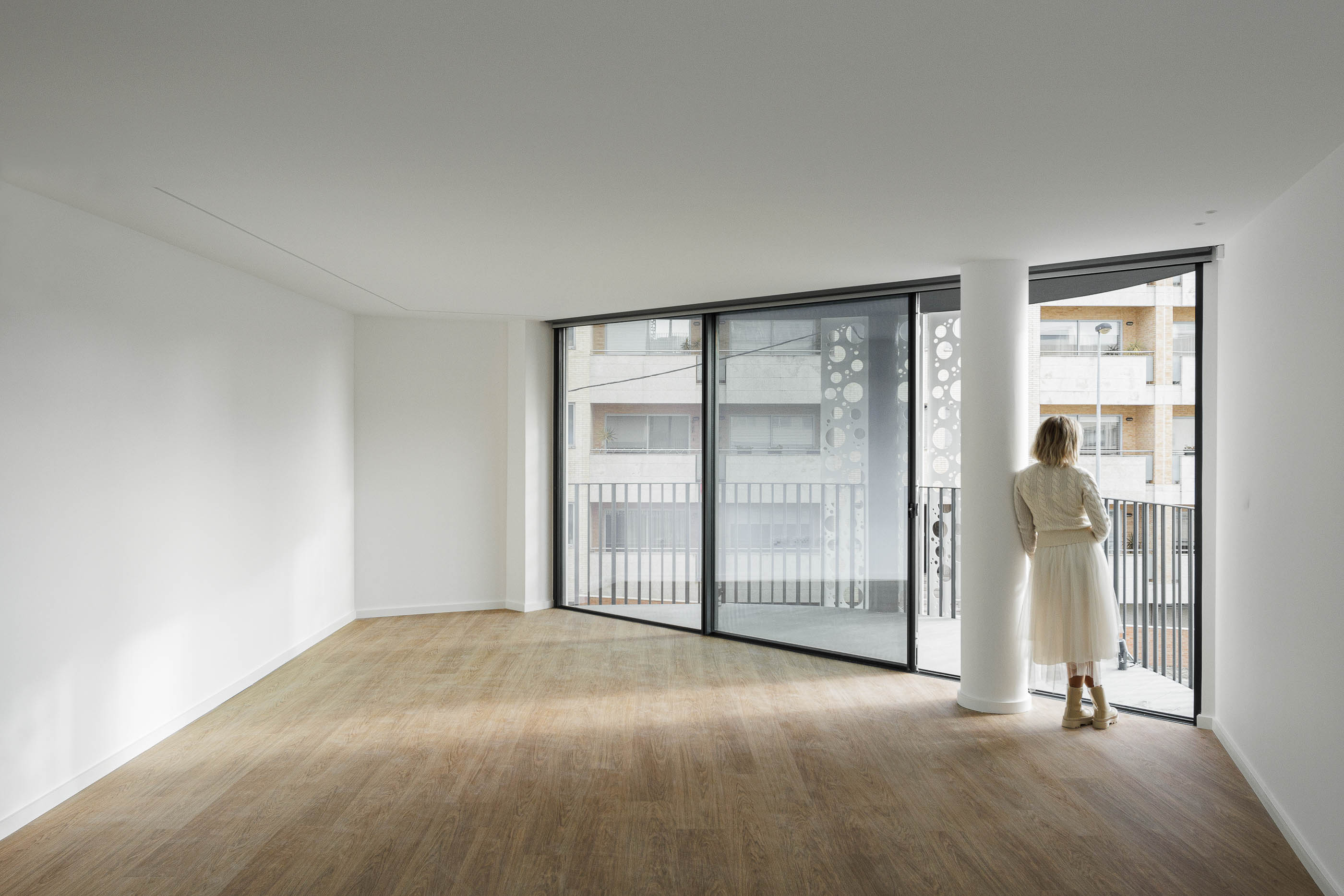 Sala de estar: Espaço amplo com pavimento em madeira, grandes janelas e acesso à varanda com elementos decorativos. — Edifício Sense 100 arquitetura Sandra Micaela Casinha Atelier em Rio Tinto