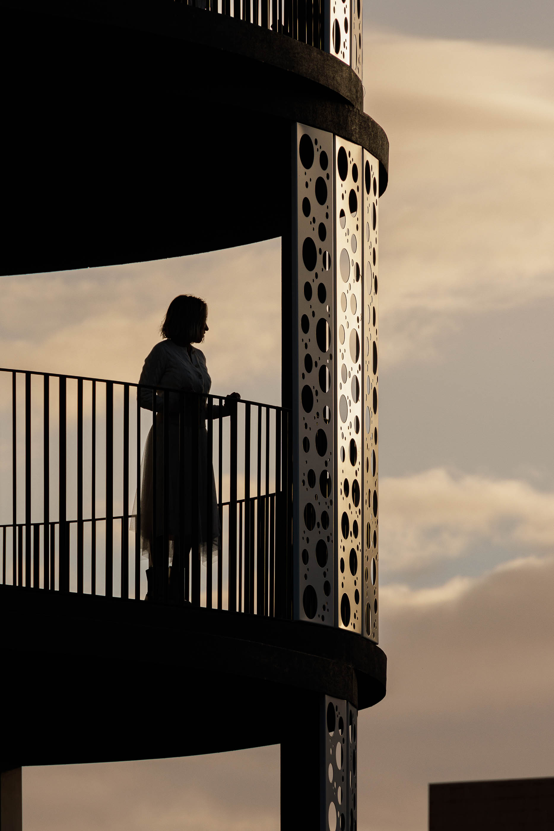 Varanda: Silhueta de uma pessoa na varanda ao pôr do sol, com estrutura perfurada e guarda-corpo em metal preto. — Edifício Sense 100 arquitetura Sandra Micaela Casinha Atelier em Rio Tinto