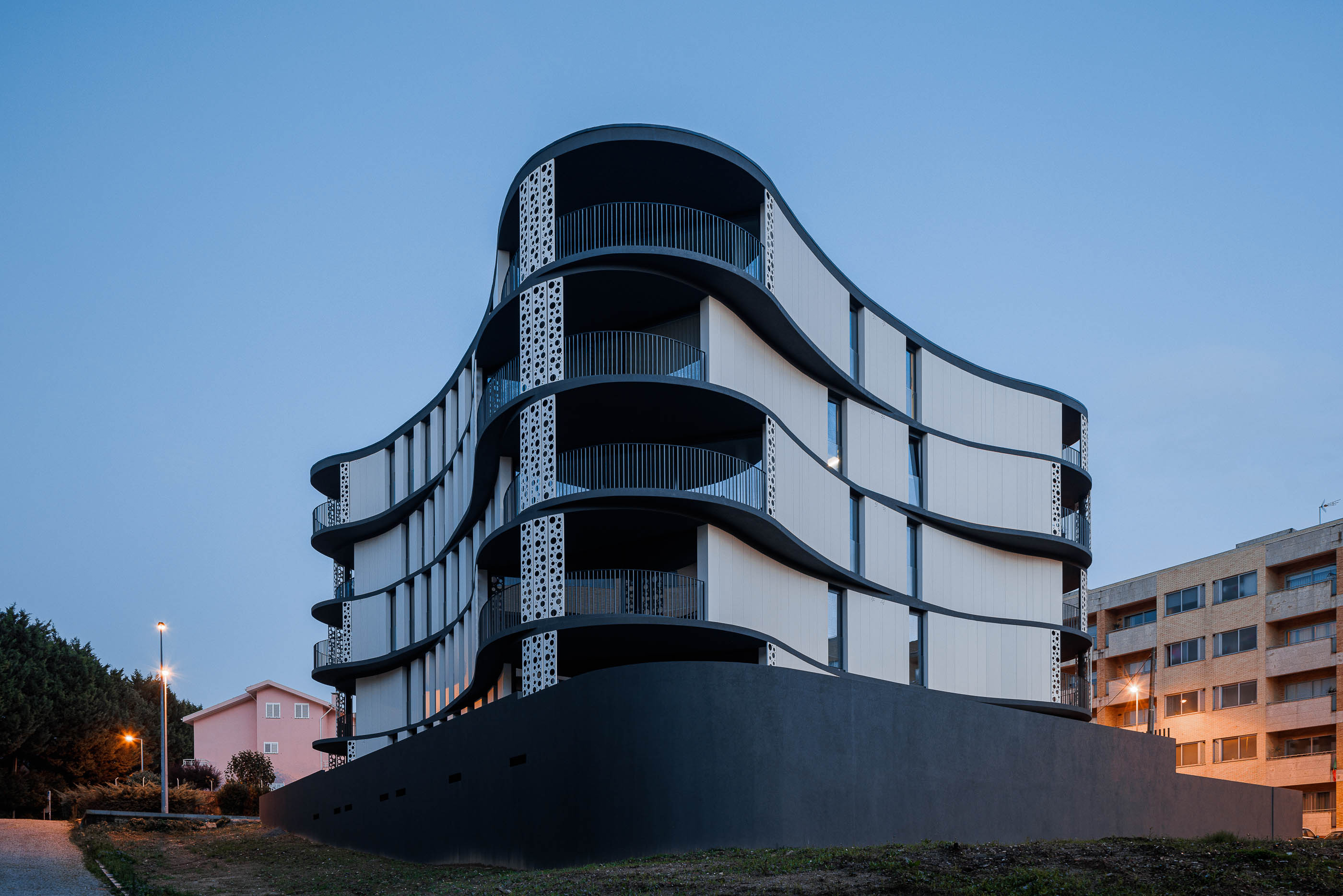 Fachada: Edifício moderno com varandas curvas e iluminação noturna, integrado na paisagem urbana. — Edifício Sense 100 arquitetura Sandra Micaela Casinha Atelier em Rio Tinto
