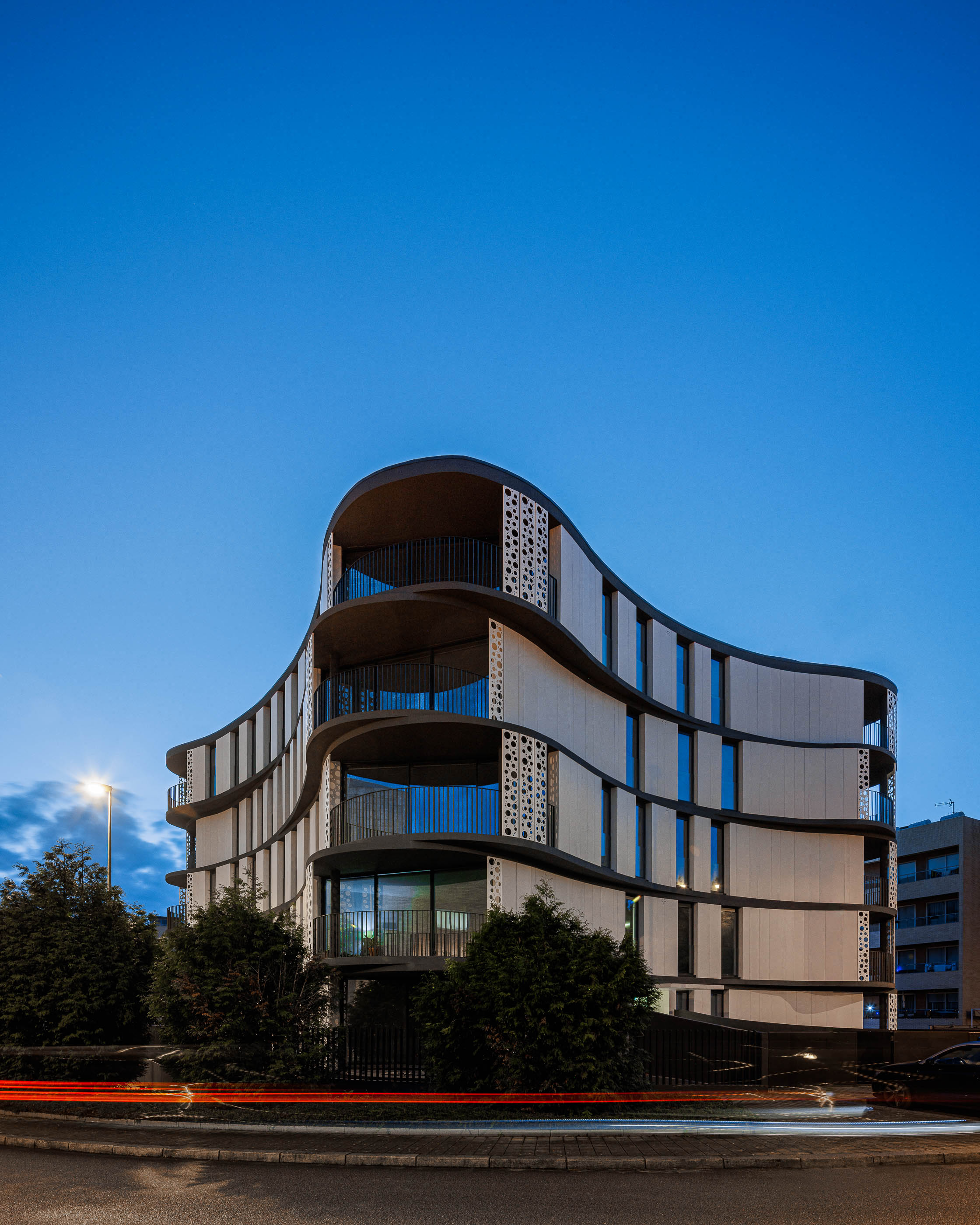 Fachada: Edifício moderno com varandas curvas e iluminação noturna, integrado na paisagem urbana. — Edifício Sense 100 arquitetura Sandra Micaela Casinha Atelier em Rio Tinto