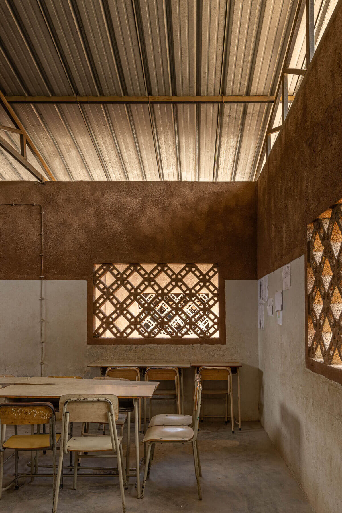 Sala de aula com paredes em tons de terra, janelas gradeadas, mesas e cadeiras de madeira sob teto metálico aparente. — Escola Kapalanga arquitetura Paulo Moreira Architectures e PARQ Arquitectos em Luanda