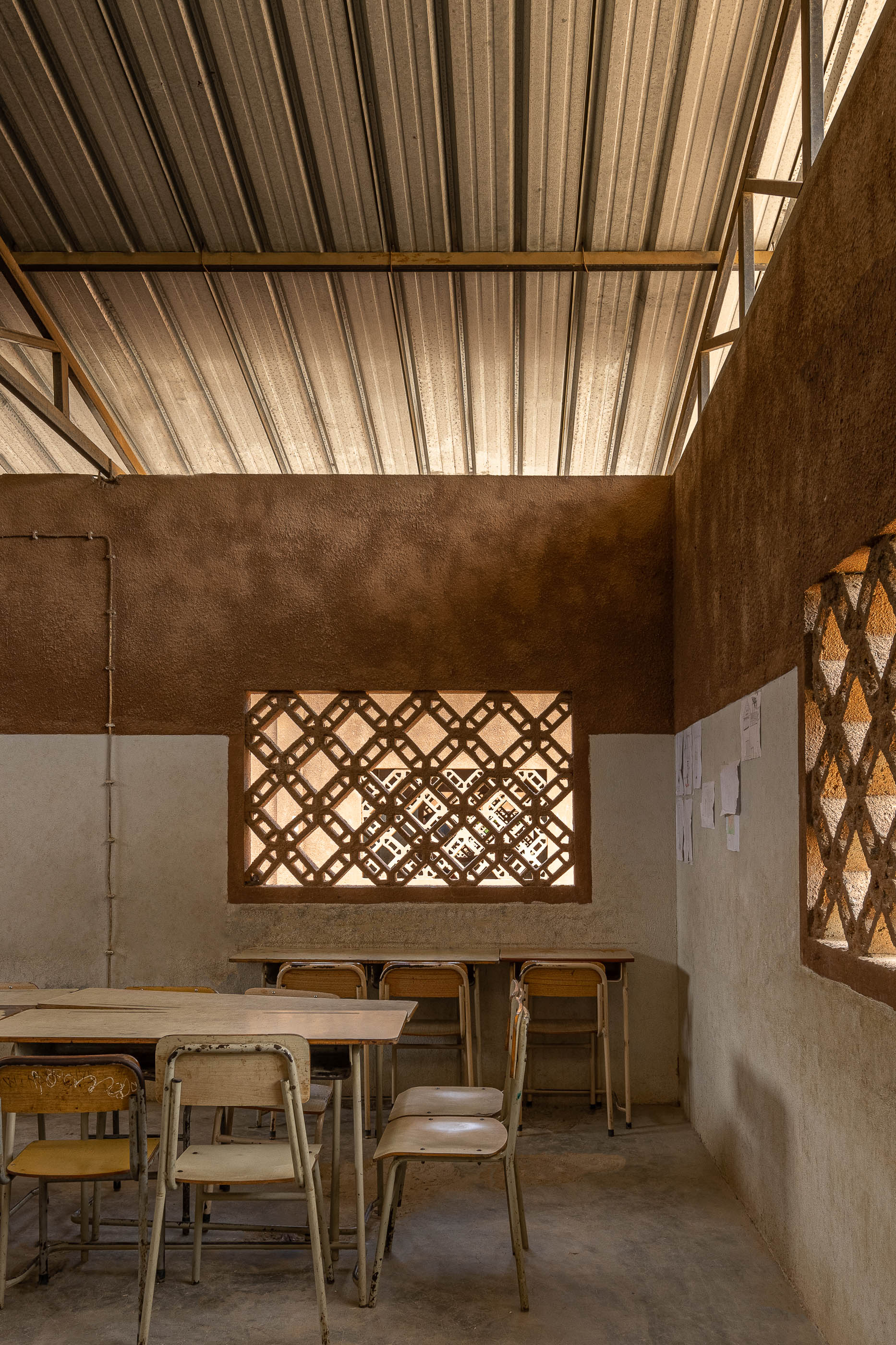Sala de aula com paredes em tons de terra, janelas gradeadas, mesas e cadeiras de madeira sob teto metálico aparente. — Escola Kapalanga arquitetura Paulo Moreira Architectures e PARQ Arquitectos em Luanda
