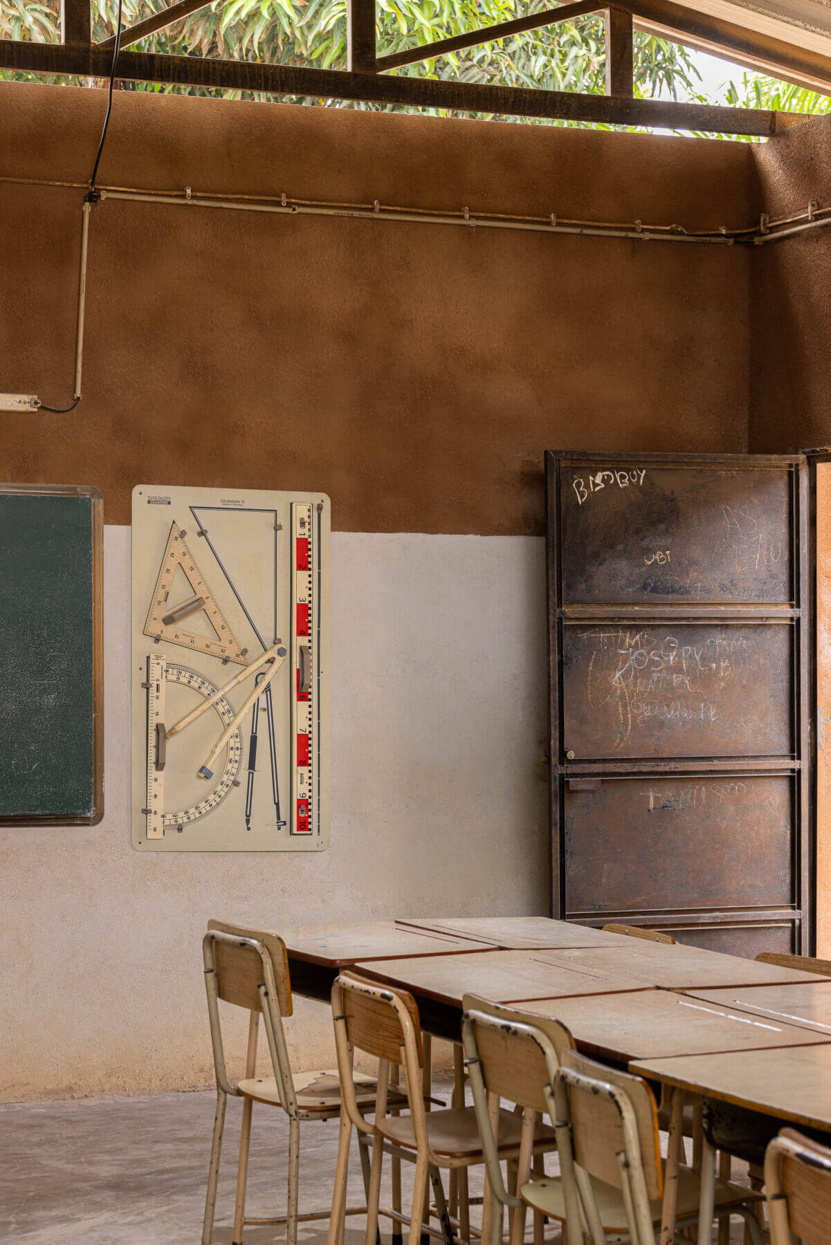 Sala de aula com quadro negro, material didático e carteiras de madeira, sob teto metálico e paredes em dois tons. — Escola Kapalanga arquitetura Paulo Moreira Architectures e PARQ Arquitectos em Luanda