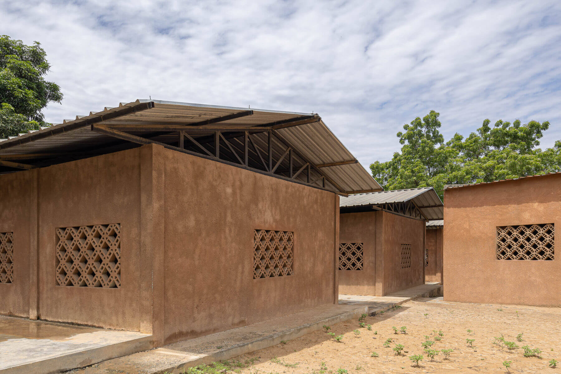 Exterior da escola com edifícios térreos de paredes texturadas em tons de terra e janelas gradeadas, sob telhados metálicos. — Escola Kapalanga arquitetura Paulo Moreira Architectures e PARQ Arquitectos em Luanda