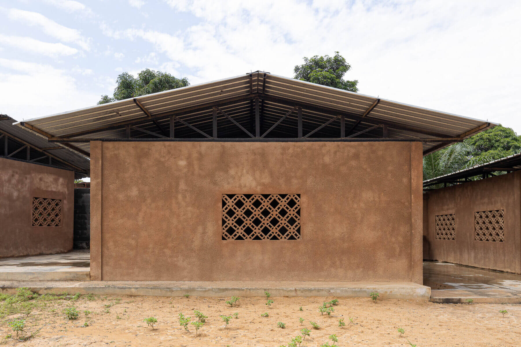 Fachada de um edifício escolar com paredes texturadas em tons de terra, janelas gradeadas e telhado metálico. — Escola Kapalanga arquitetura Paulo Moreira Architectures e PARQ Arquitectos em Luanda