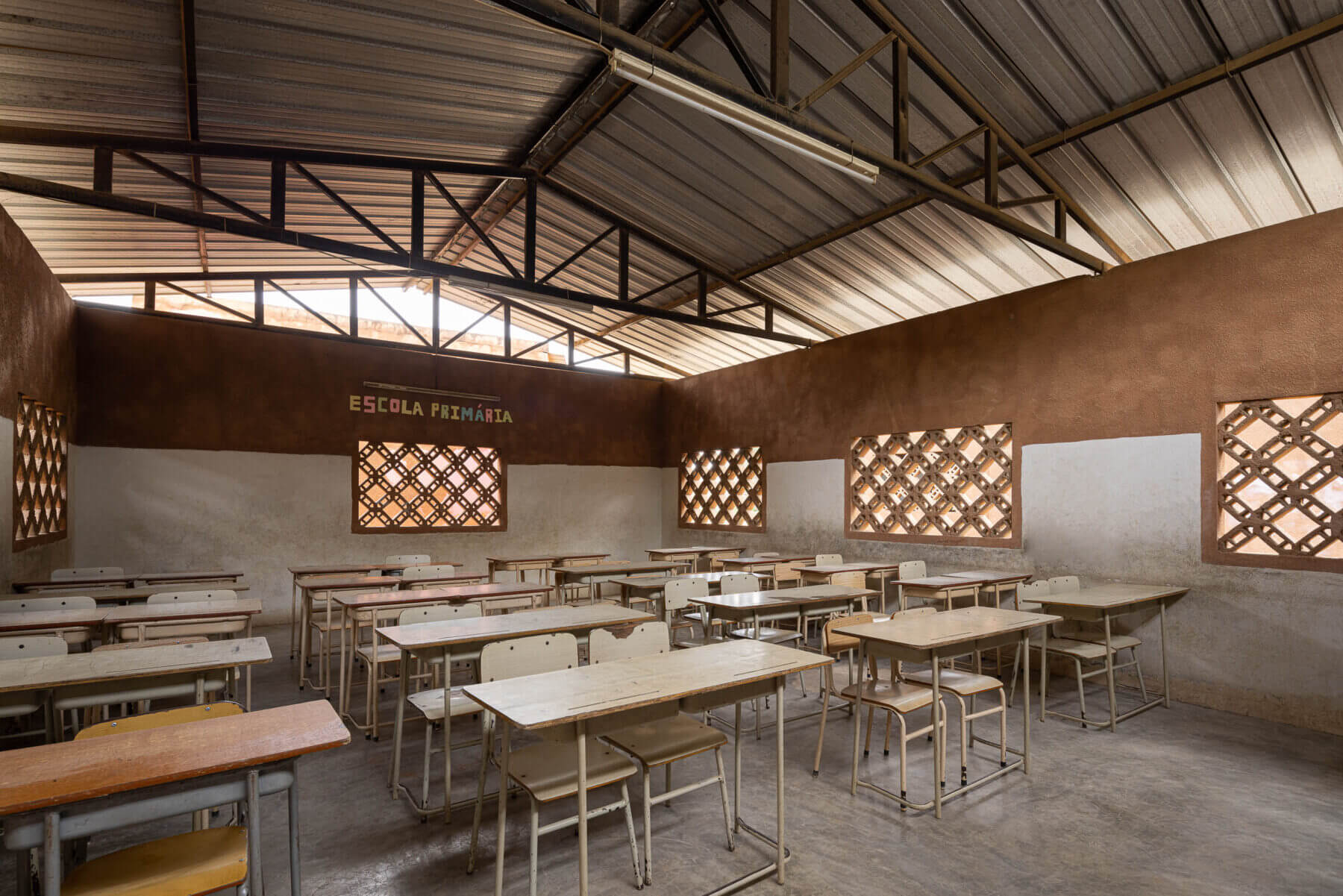 Sala de aula ampla com carteiras de madeira, janelas gradeadas e paredes em dois tons, sob teto metálico aparente. — Escola Kapalanga arquitetura Paulo Moreira Architectures e PARQ Arquitectos em Luanda