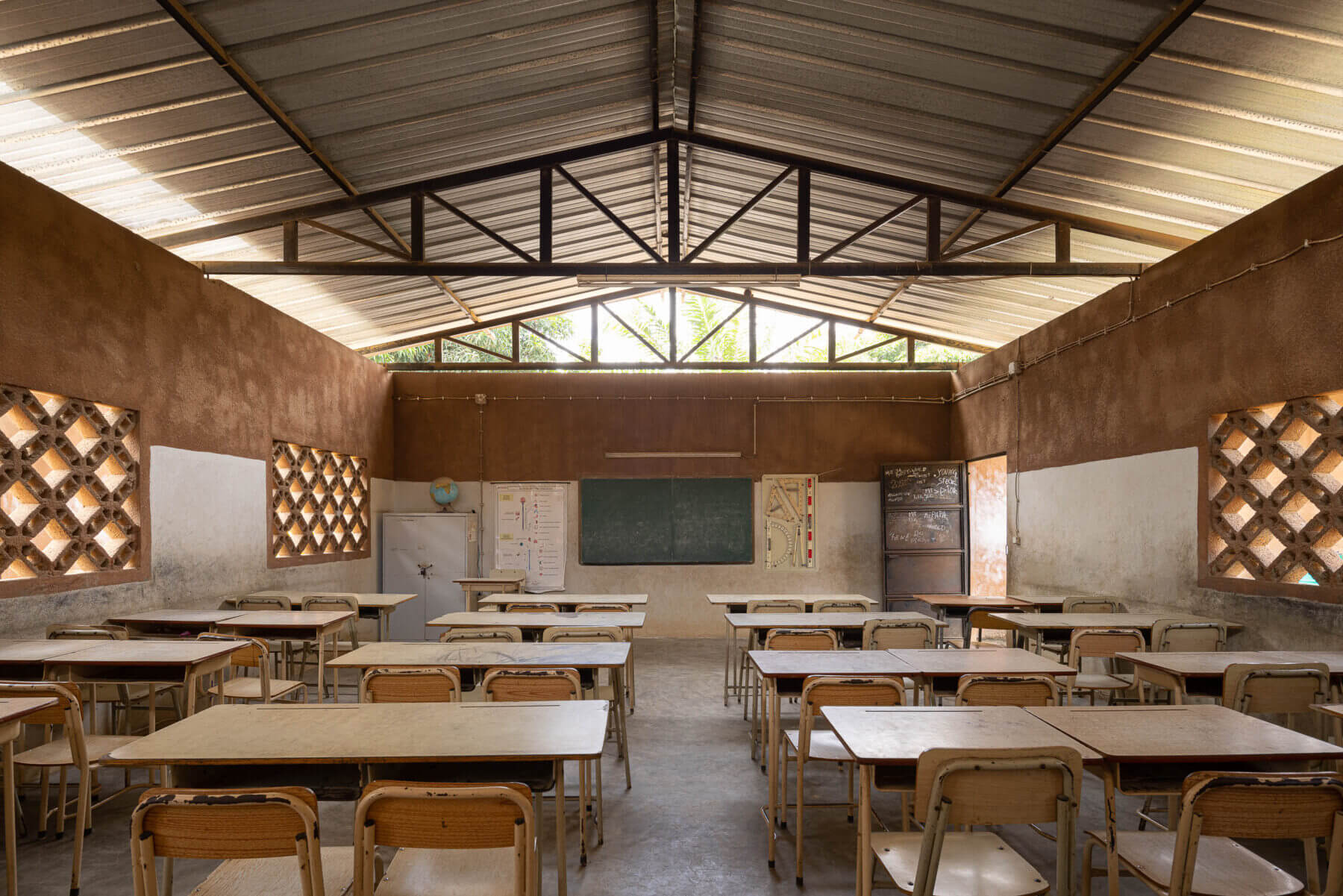 Sala de aula com carteiras de madeira, quadro negro e janelas gradeadas, sob teto metálico e paredes em dois tons. — Escola Kapalanga arquitetura Paulo Moreira Architectures e PARQ Arquitectos em Luanda