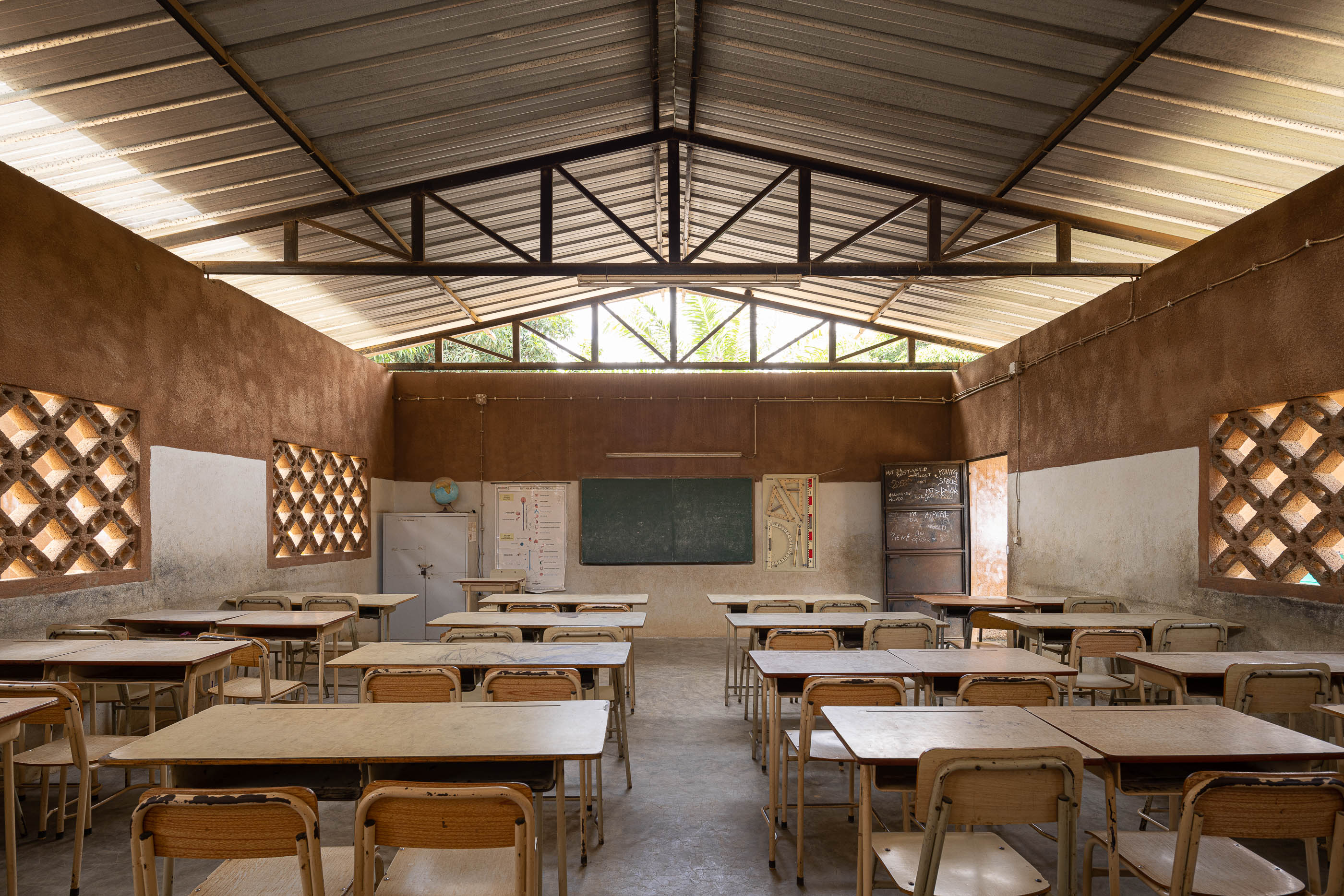 Sala de aula com carteiras de madeira, quadro negro e janelas gradeadas, sob teto metálico e paredes em dois tons. — Escola Kapalanga arquitetura Paulo Moreira Architectures e PARQ Arquitectos em Luanda
