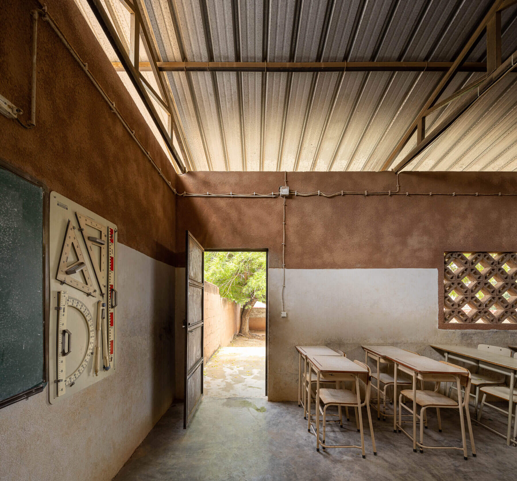 Sala de aula com porta aberta para o exterior, quadro negro e carteiras de madeira, sob teto metálico aparente. — Escola Kapalanga arquitetura Paulo Moreira Architectures e PARQ Arquitectos em Luanda