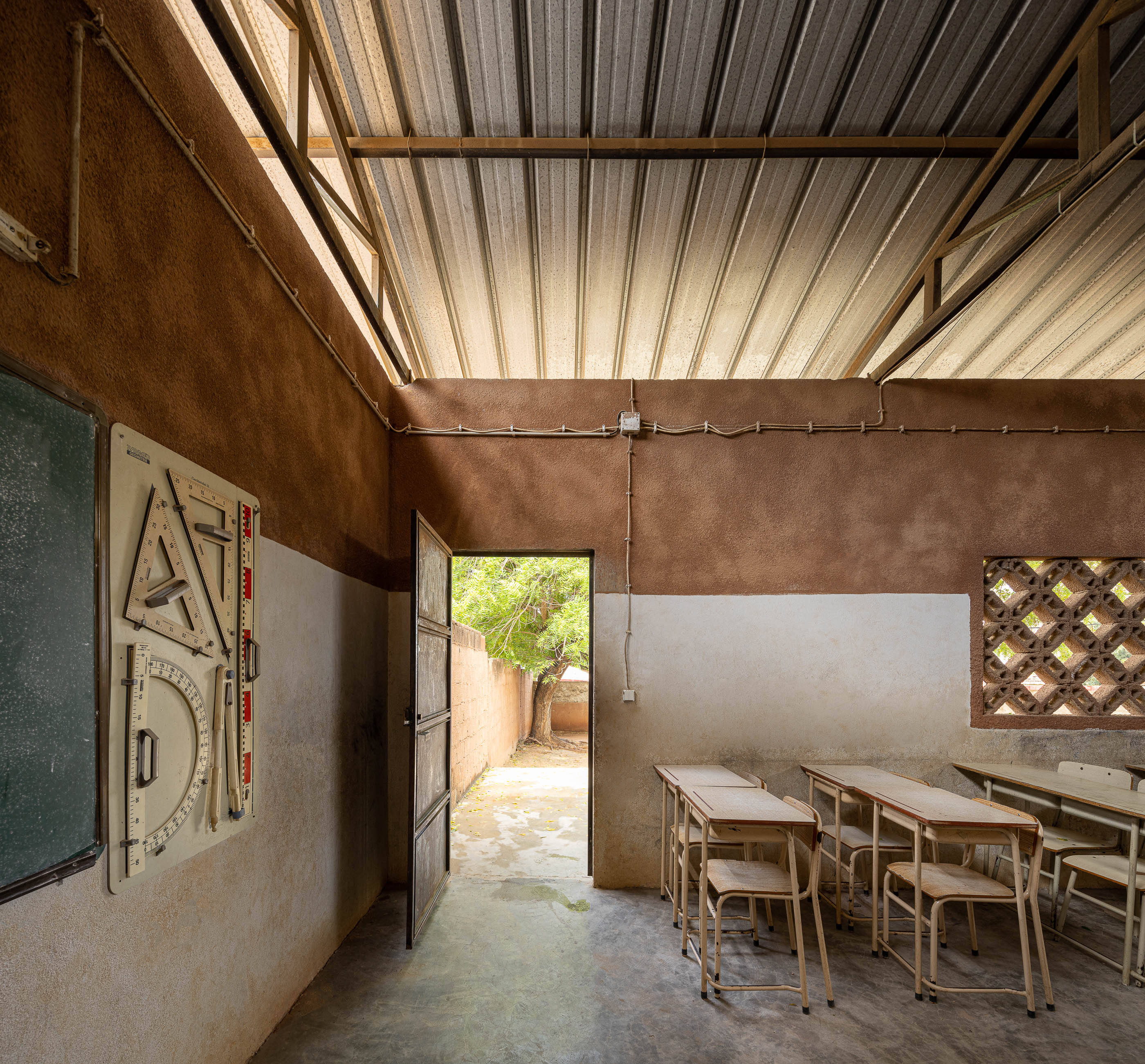 Sala de aula com porta aberta para o exterior, quadro negro e carteiras de madeira, sob teto metálico aparente. — Escola Kapalanga arquitetura Paulo Moreira Architectures e PARQ Arquitectos em Luanda